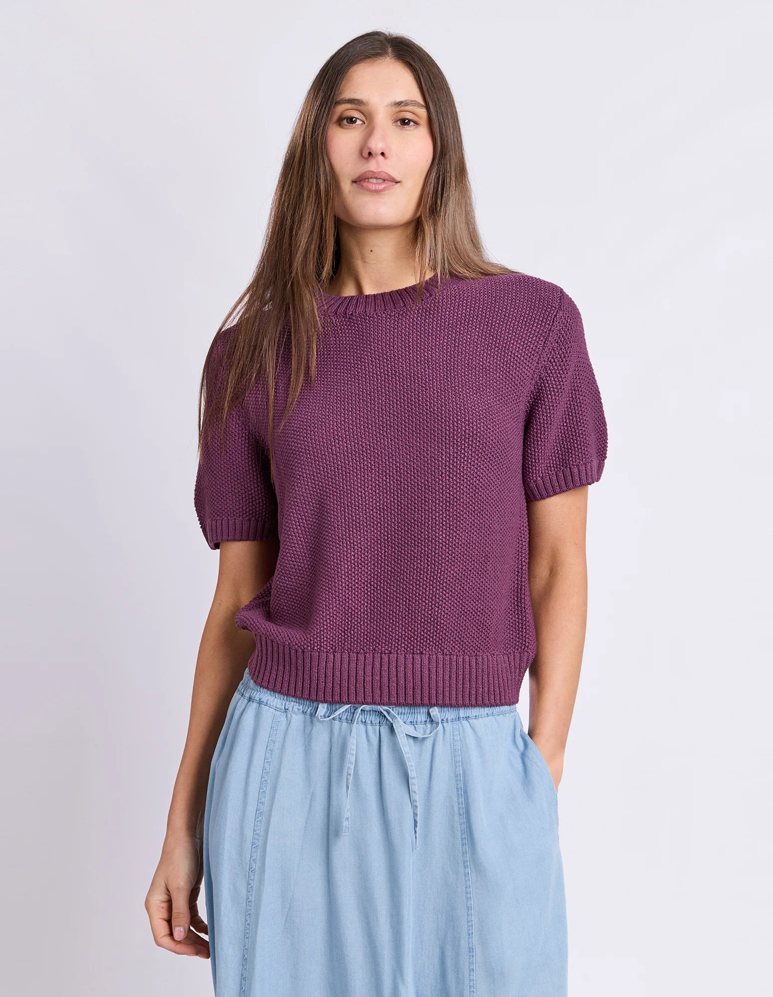 Image of A woman modeling a Foxwood short-sleeve knit sweater in a muted plum purple color, paired with light blue drawstring pants, against a plain light gray studio background.