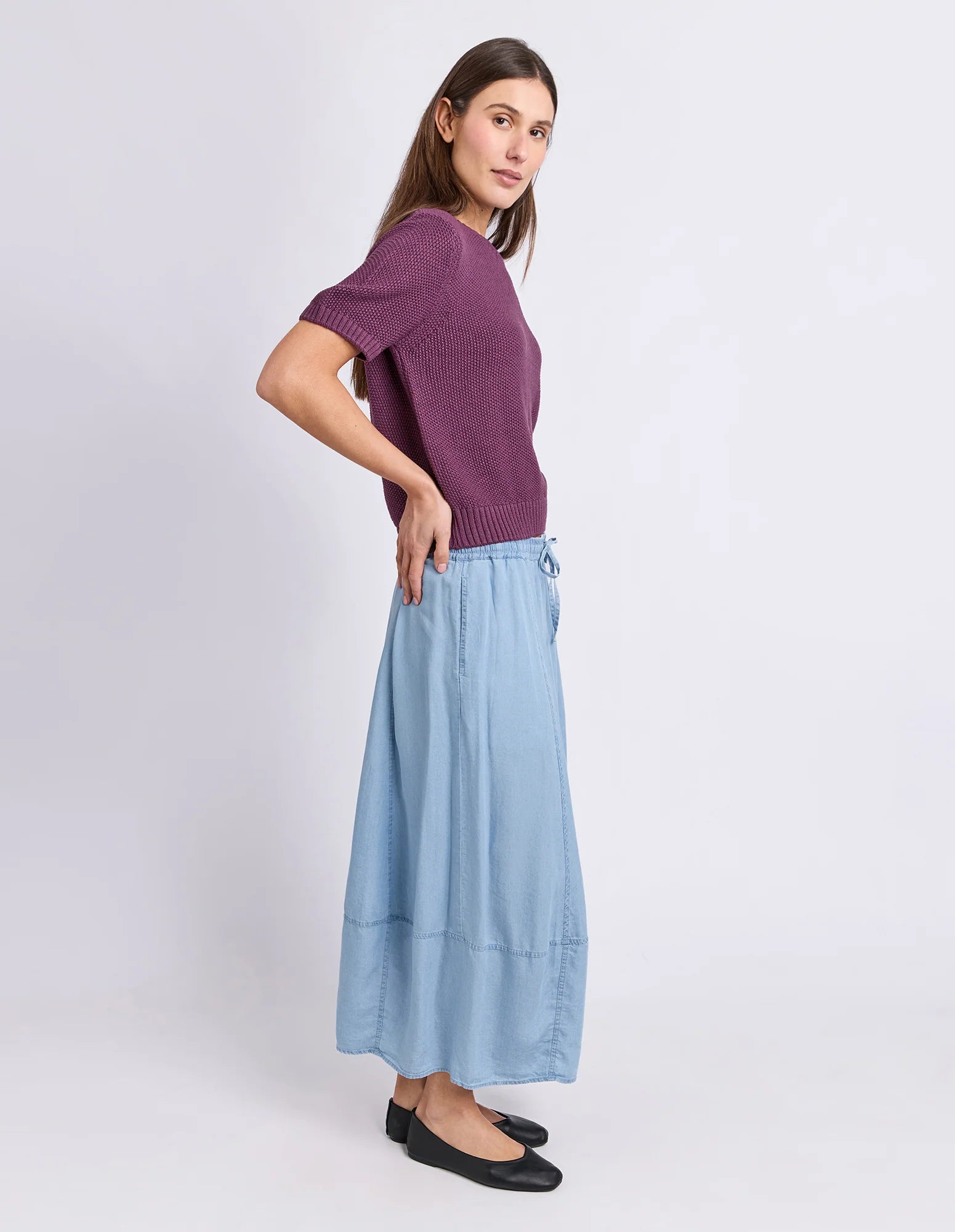 Image of A woman modeling a Foxwood short-sleeve purple knit top and a light blue wide-leg skirt, paired with black flats, against a plain light gray studio background.