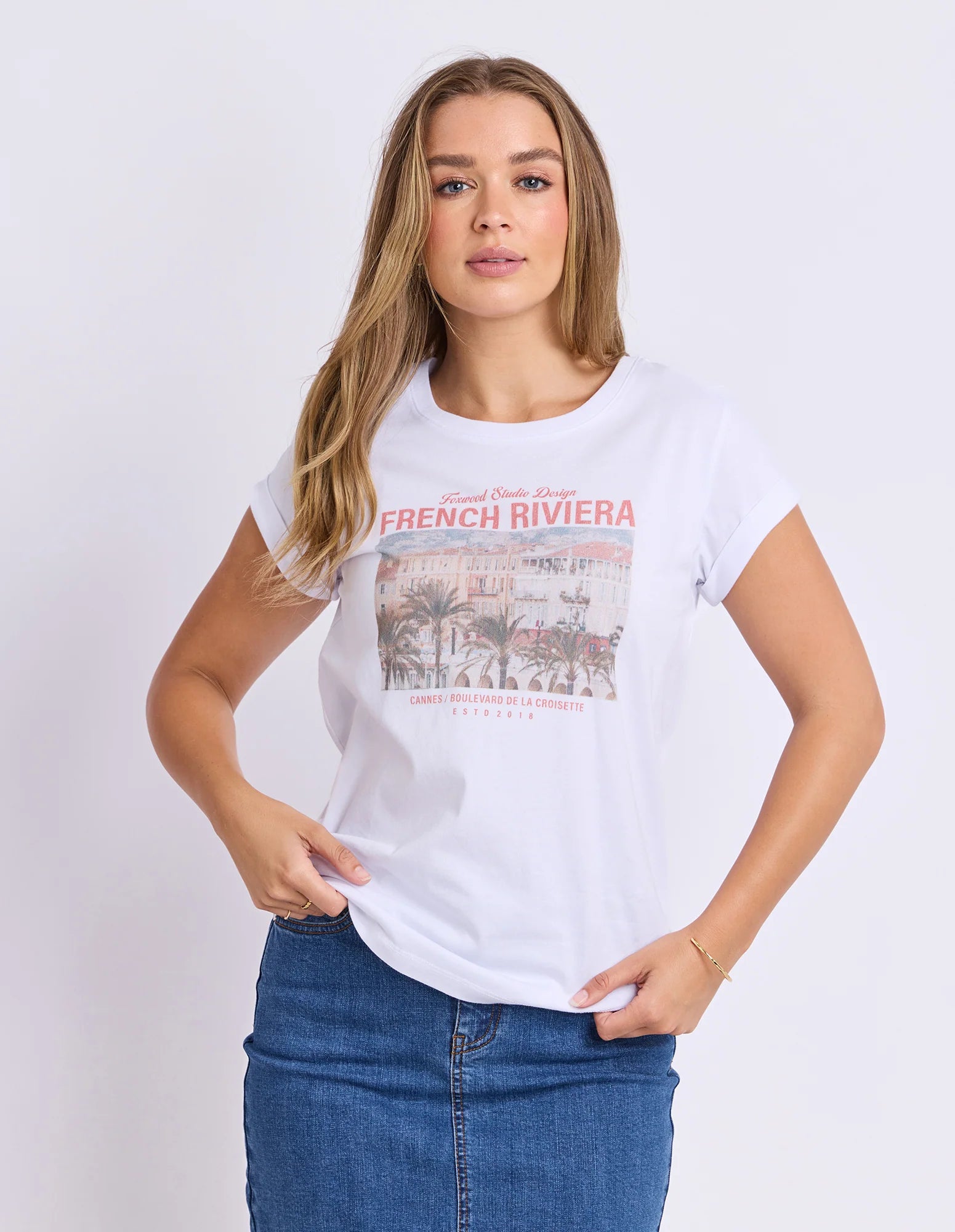 Image of A woman modeling a Foxwood white short-sleeve graphic t-shirt with a vintage-style 'French Riviera' print on the front, paired with a blue denim skirt, against a plain white studio background.