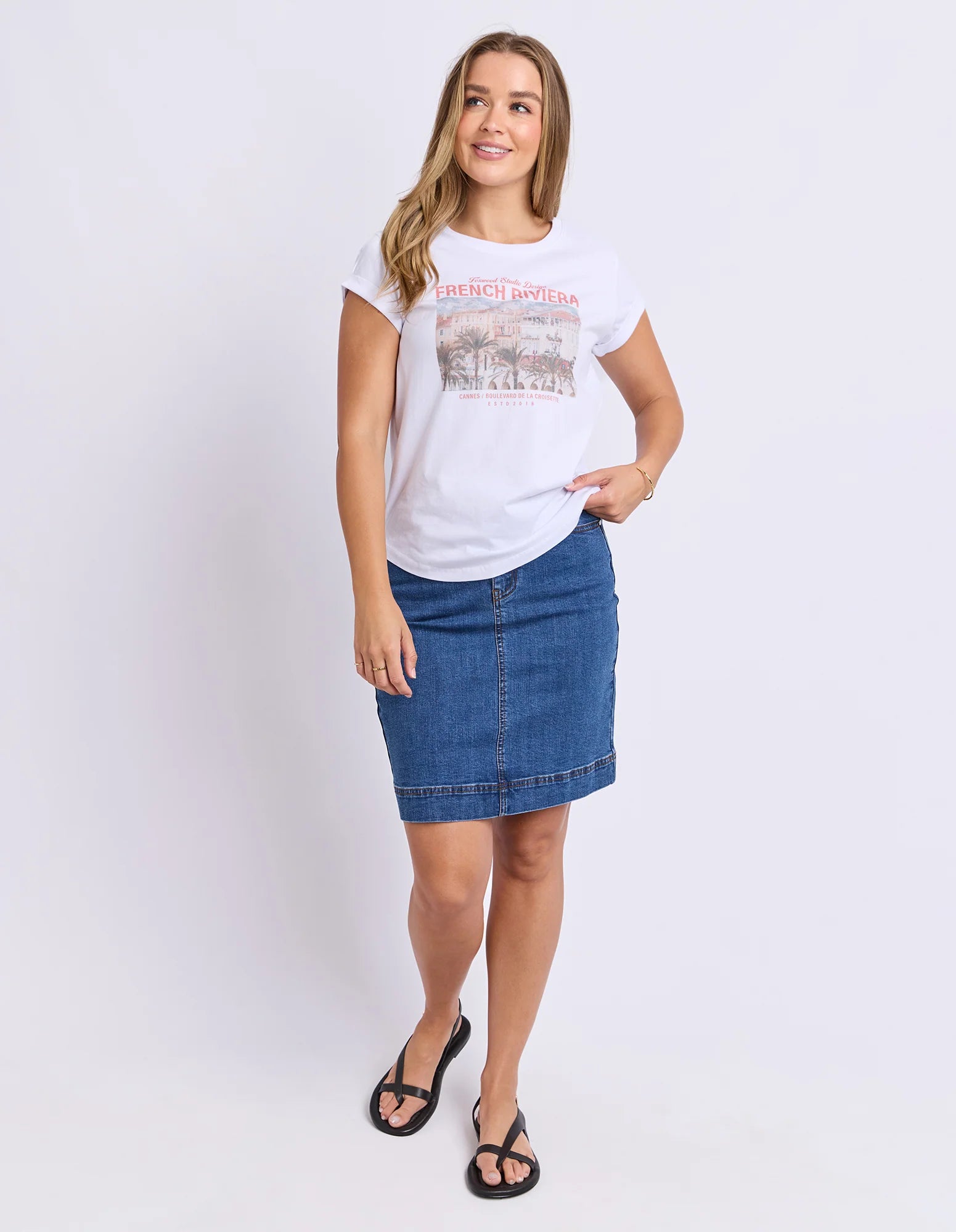Image of A woman modeling a Foxwood white graphic t-shirt and a blue denim pencil skirt, paired with black thong sandals, against a plain white studio background.