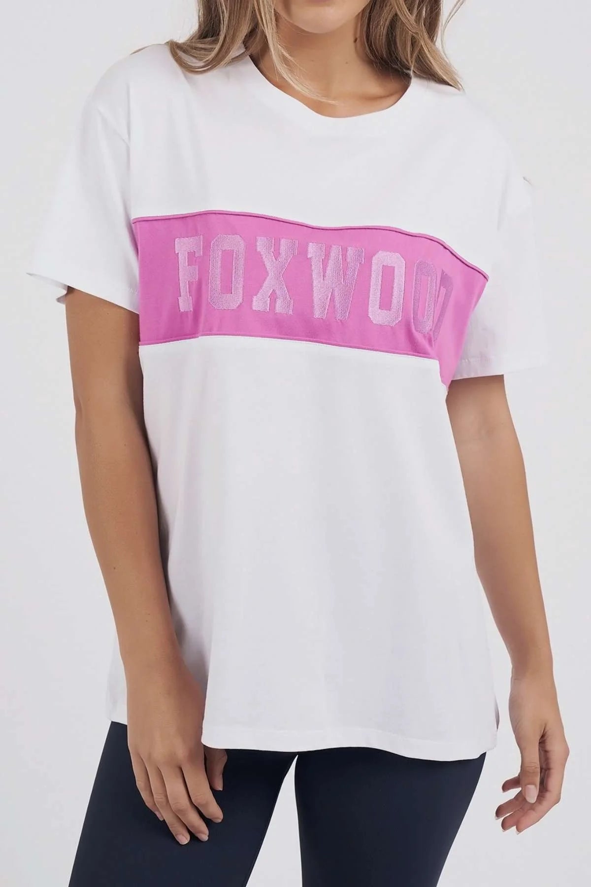 Image of A woman wearing a Foxwood white short-sleeve T-shirt with a wide pink horizontal band across the chest featuring the word 'FOXWOOL' in light pink lettering, paired with dark leggings against a plain light background.
