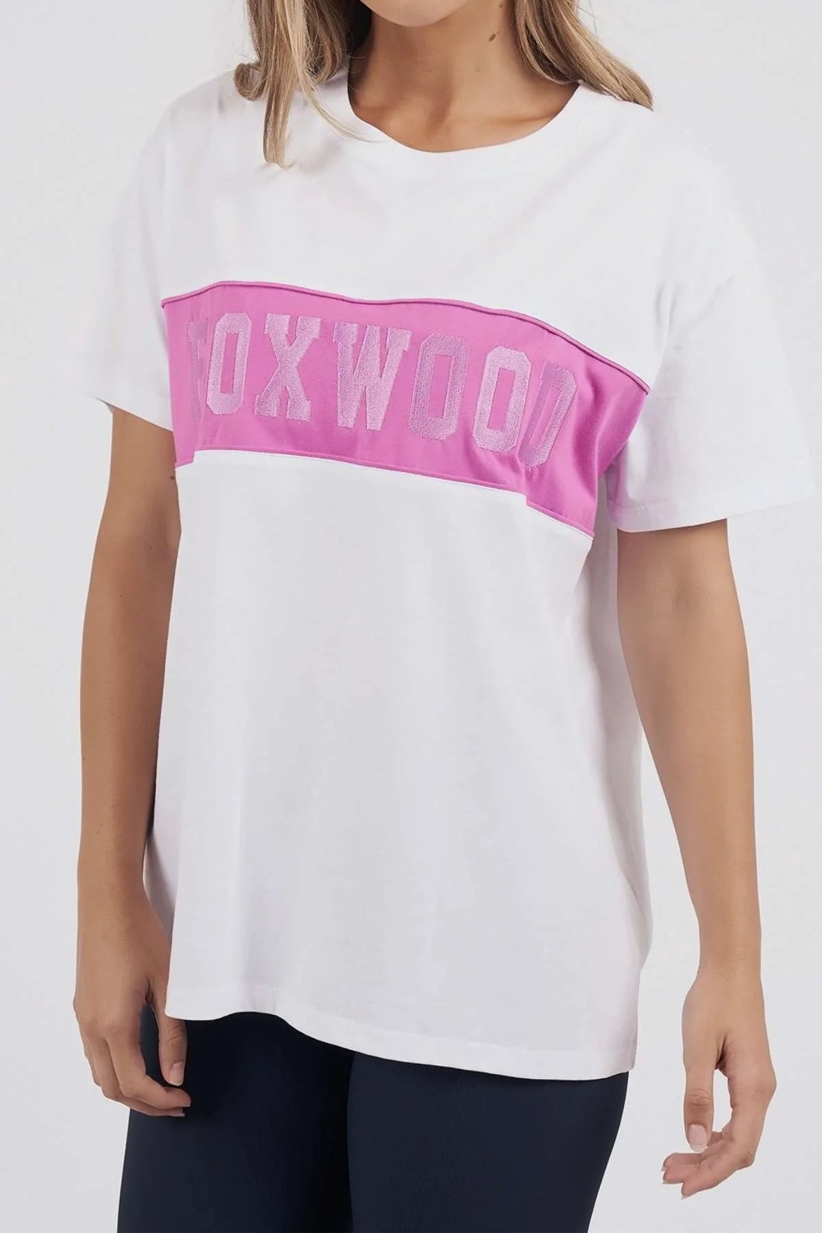 Image of A woman wearing a Foxwood white short-sleeve t-shirt with a bright pink horizontal chest stripe featuring the word 'FOXWOOD' in light pink letters, paired with dark leggings against a plain white background.