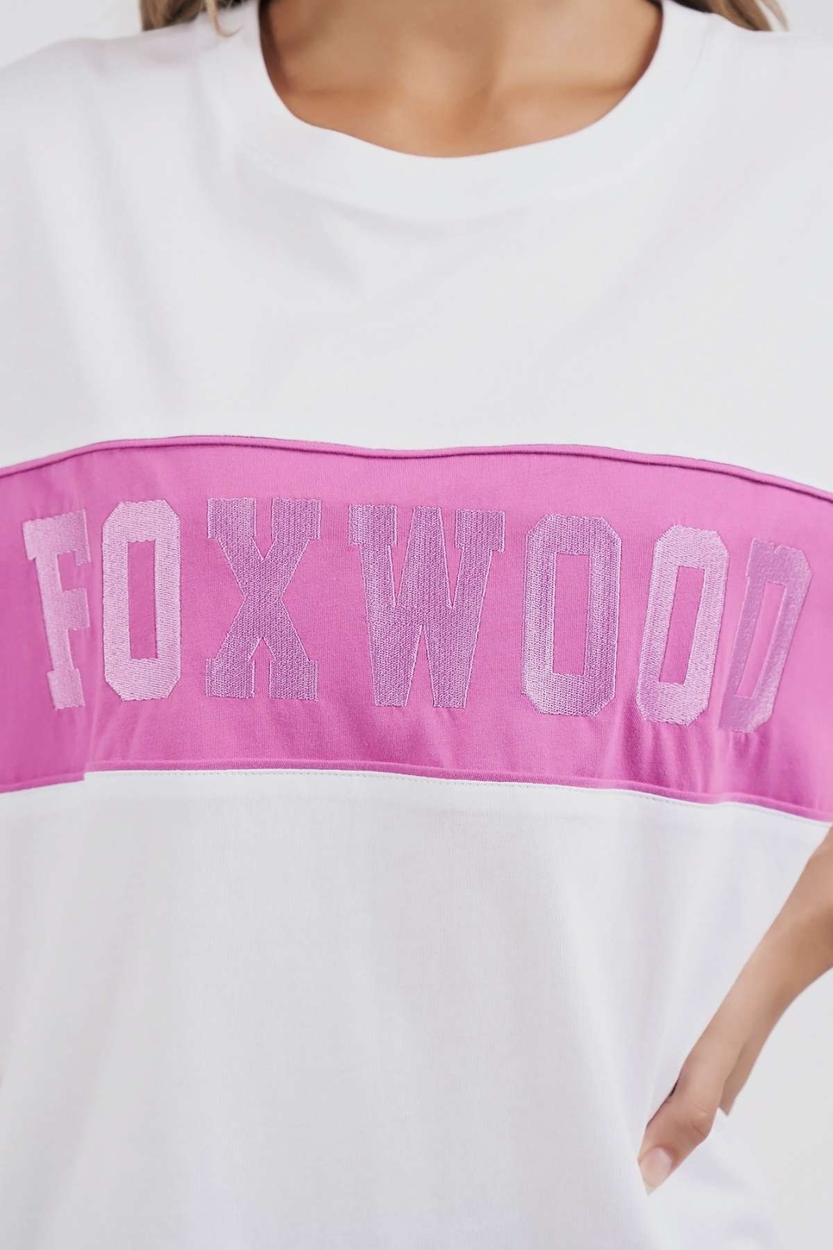 Image of A close-up of a Foxwood white crewneck tshirt featuring a wide pink horizontal stripe across the chest with the word 'FOXWOOD' embroidered in light pink letters.
