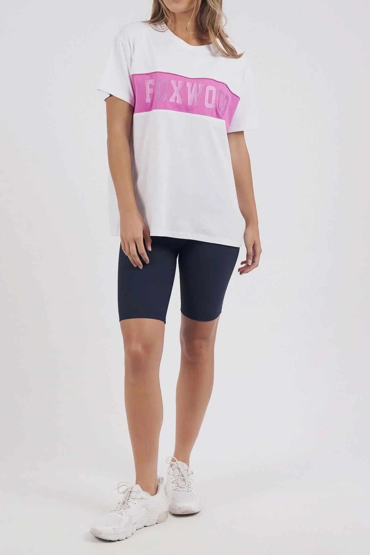 Image of A woman modeling a Foxwood white short-sleeve t-shirt with a pink horizontal stripe across the chest featuring the word 'EXWOO' in white, paired with dark navy bike shorts and white sneakers, against a plain white studio background.
