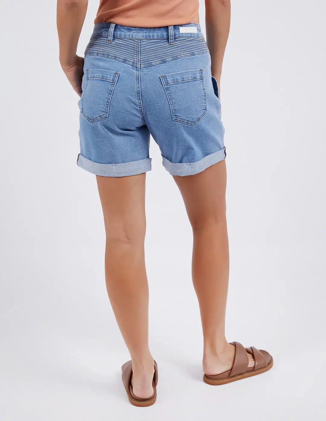 Image of A woman shown from the back wearing Foxwood light blue denim shorts with rolled hems, a peach top, and brown slide sandals against a plain light gray studio background.