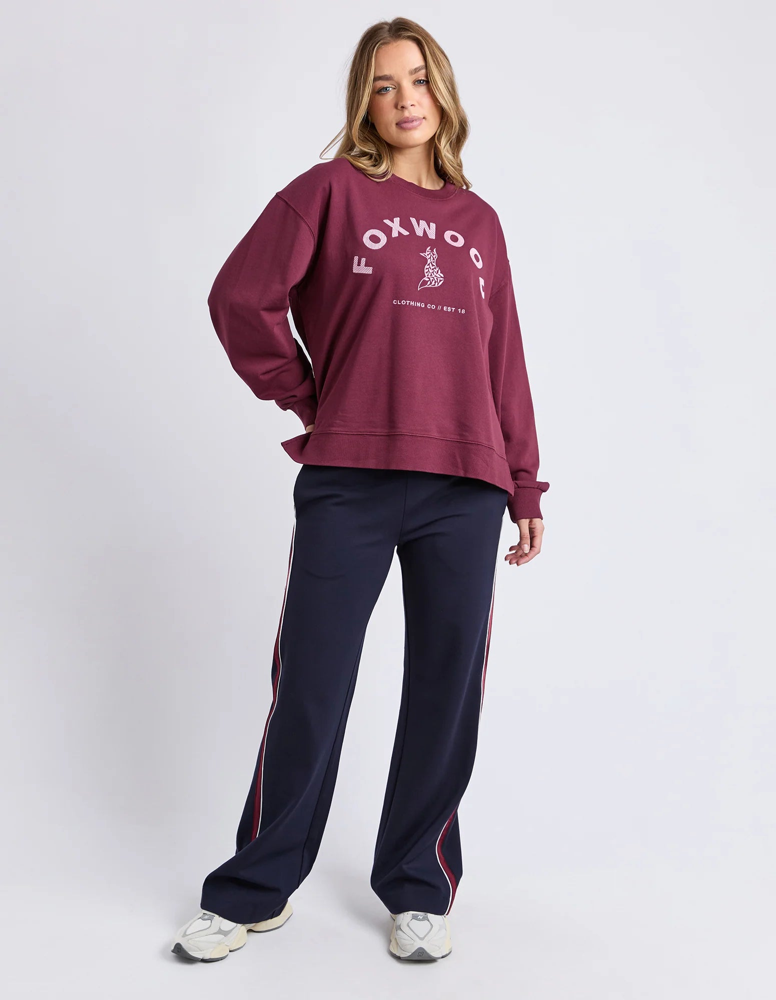 Image of Woman wearing a Foxwood maroon sweatshirt with 'Foxwood' logo and navy tracksuit pants on a white background