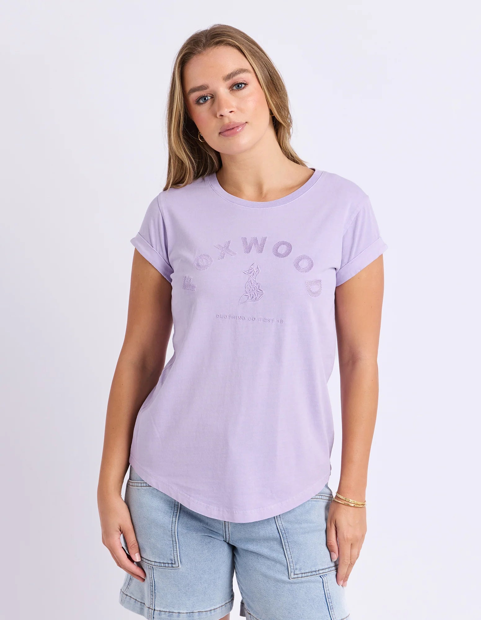 Image of A woman wearing a Foxwood light purple short-sleeve t-shirt with a tonal 'FOXWOOD' graphic on the front, paired with light blue denim shorts, against a plain light gray studio background.