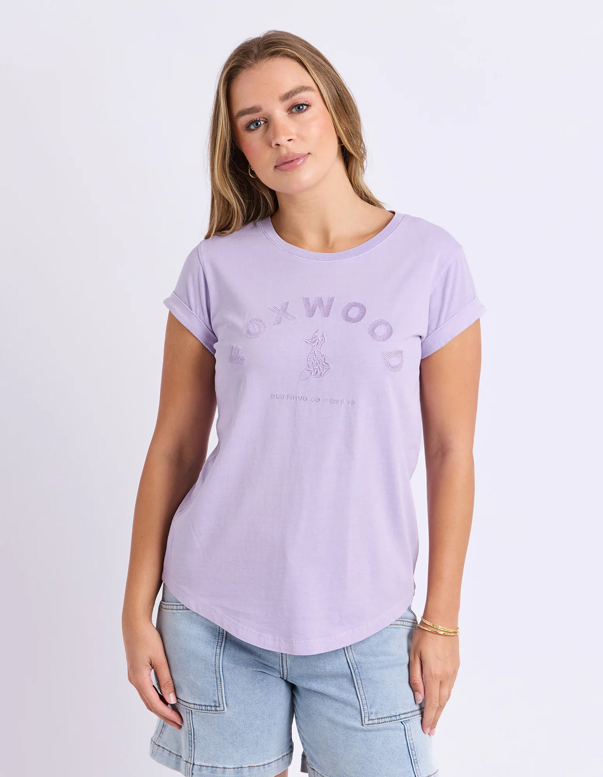 A woman wearing a Foxwood light purple short-sleeve t-shirt with a tonal 'FOXWOOD' graphic on the front, paired with light blue denim shorts, against a plain light gray studio background.