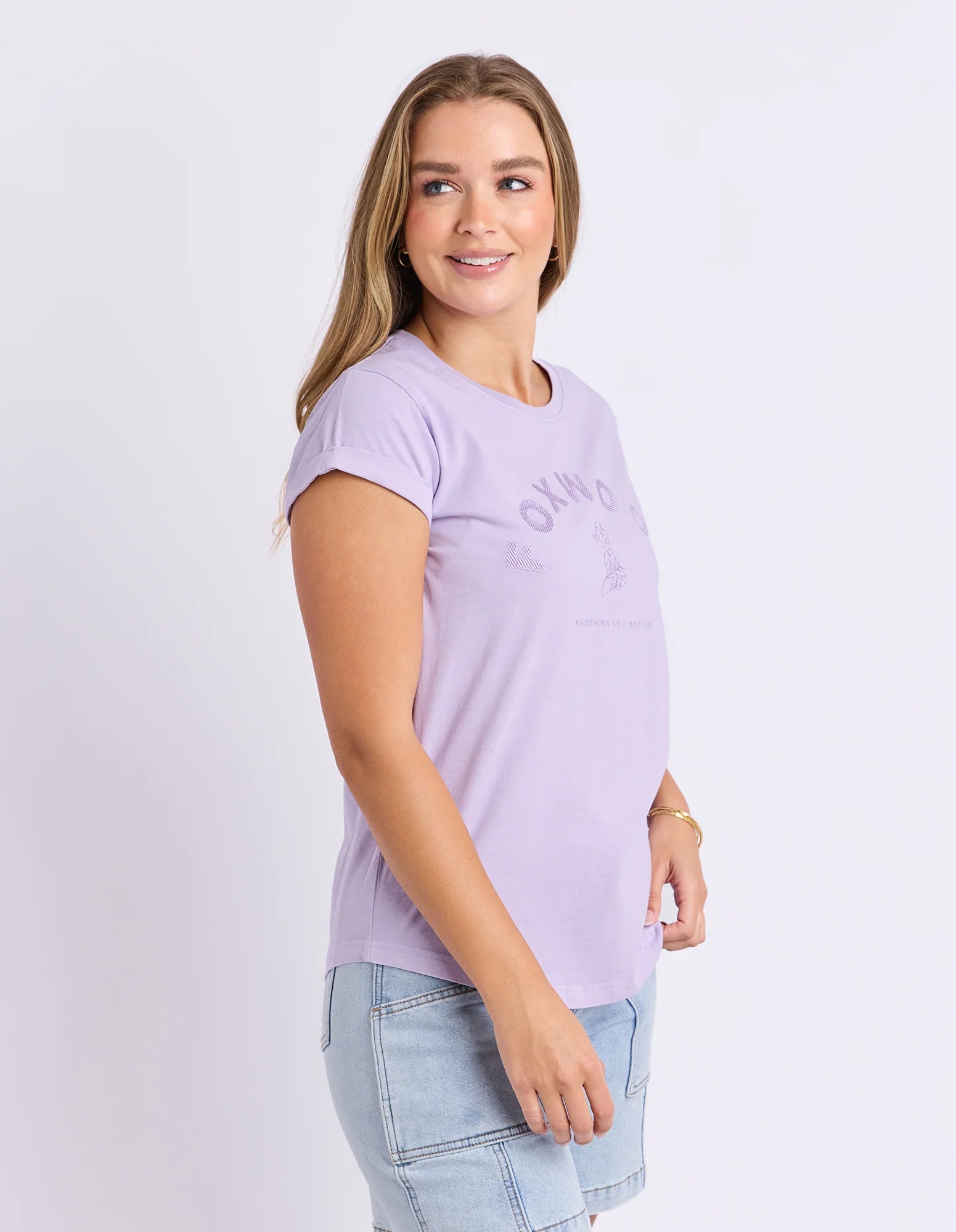 Image of A woman wearing a Foxwood light purple short-sleeve graphic t-shirt and light blue denim shorts, standing against a plain light gray studio background.