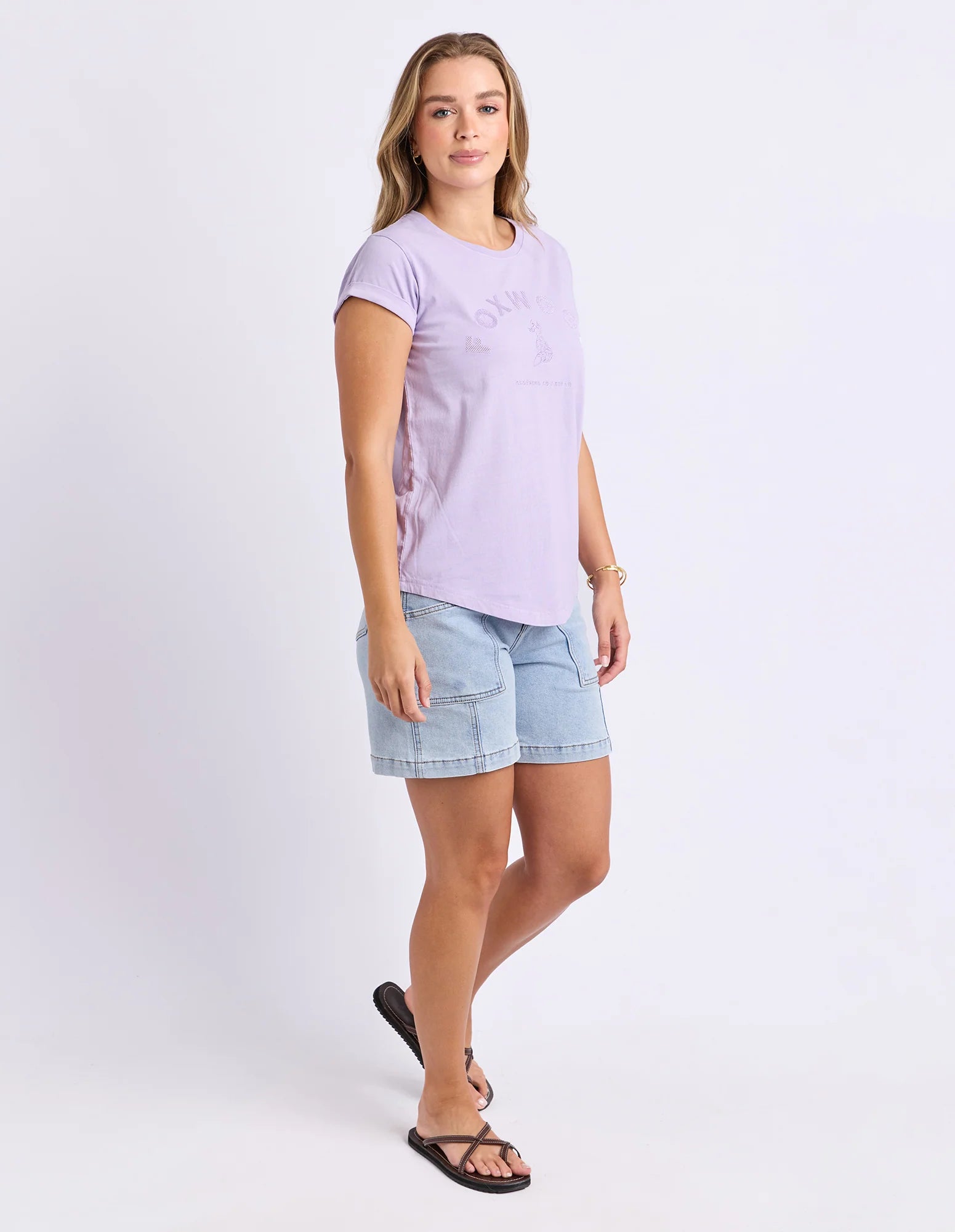 Image of A woman modeling a Foxwood light purple short-sleeve t-shirt with a tonal graphic and light blue denim shorts, paired with black sandals, against a plain white studio background.