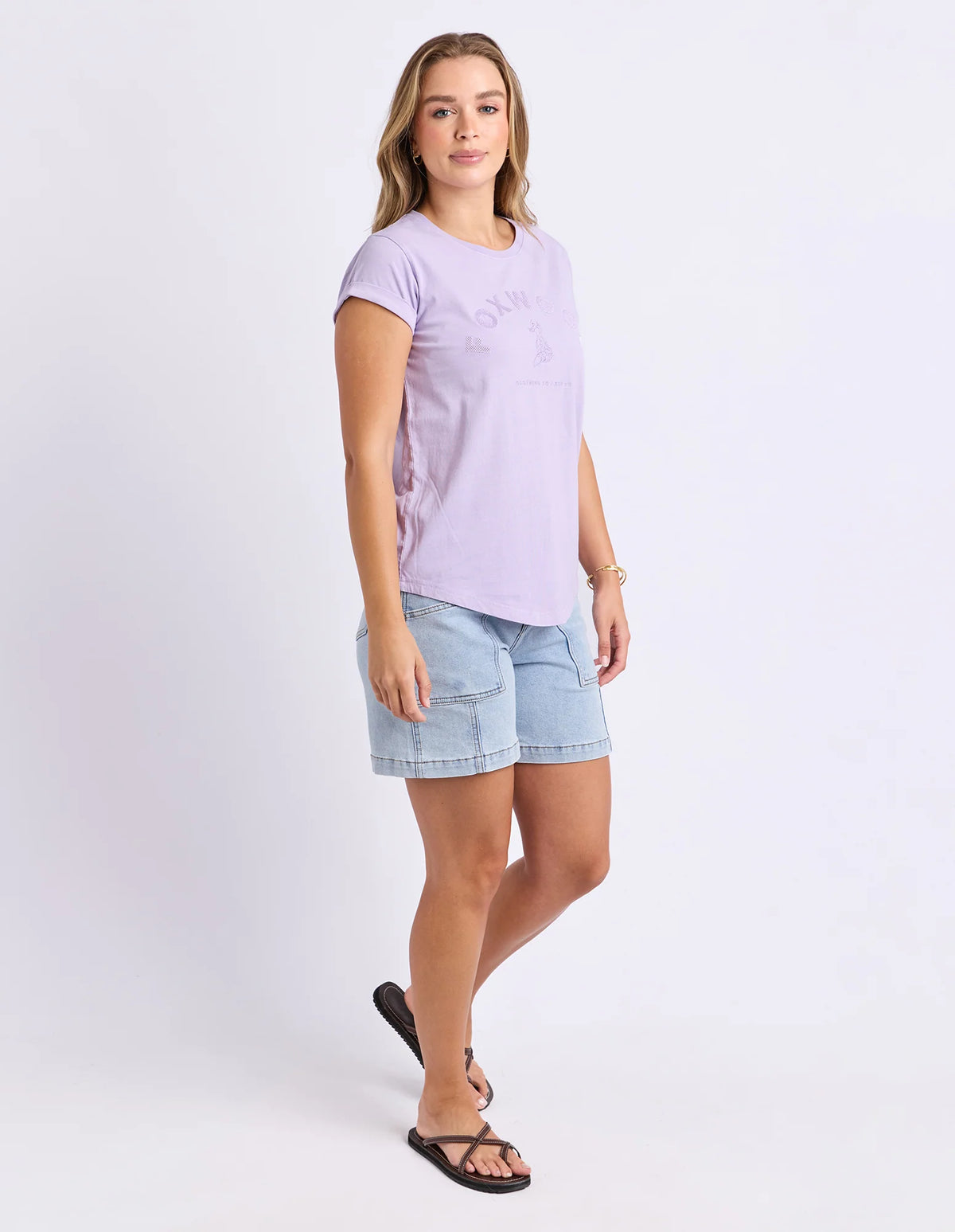 A woman modeling a Foxwood light purple short-sleeve t-shirt with a tonal graphic and light blue denim shorts, paired with black sandals, against a plain white studio background.