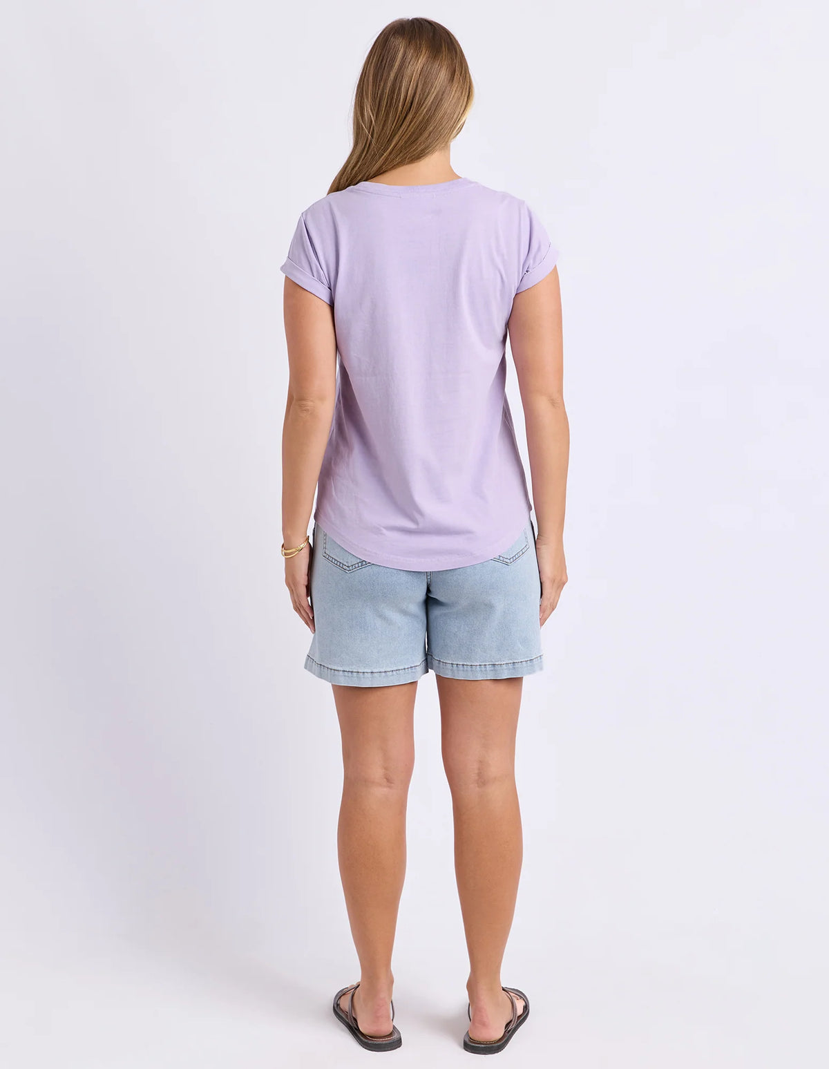 A woman viewed from behind wearing a Foxwood light purple short-sleeve t-shirt and light blue denim shorts against a plain white studio background.