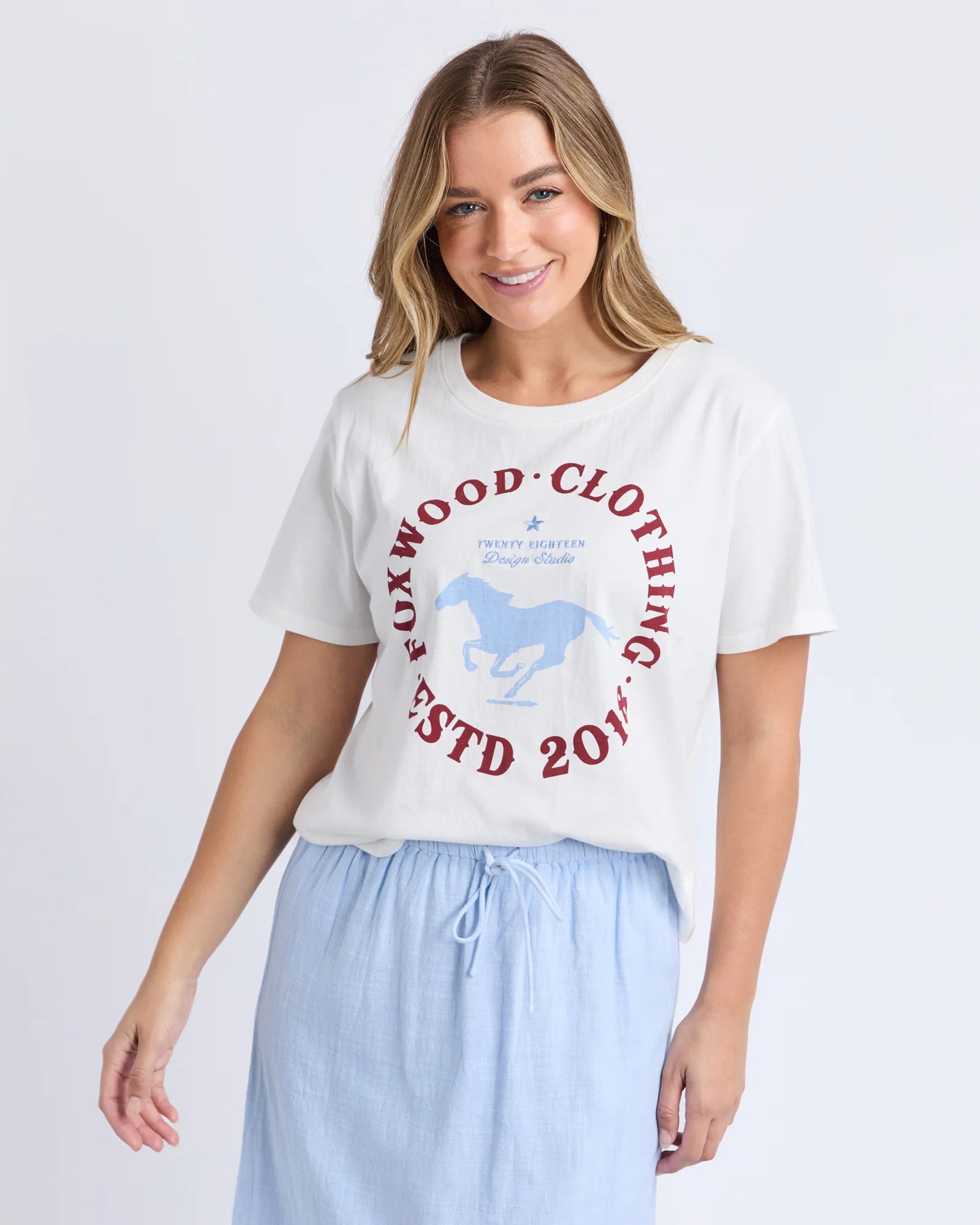 Image of A woman wearing a Foxwood white graphic t-shirt with a red and blue Foxwood Clothing logo, paired with light blue drawstring pants, against a plain light gray background.