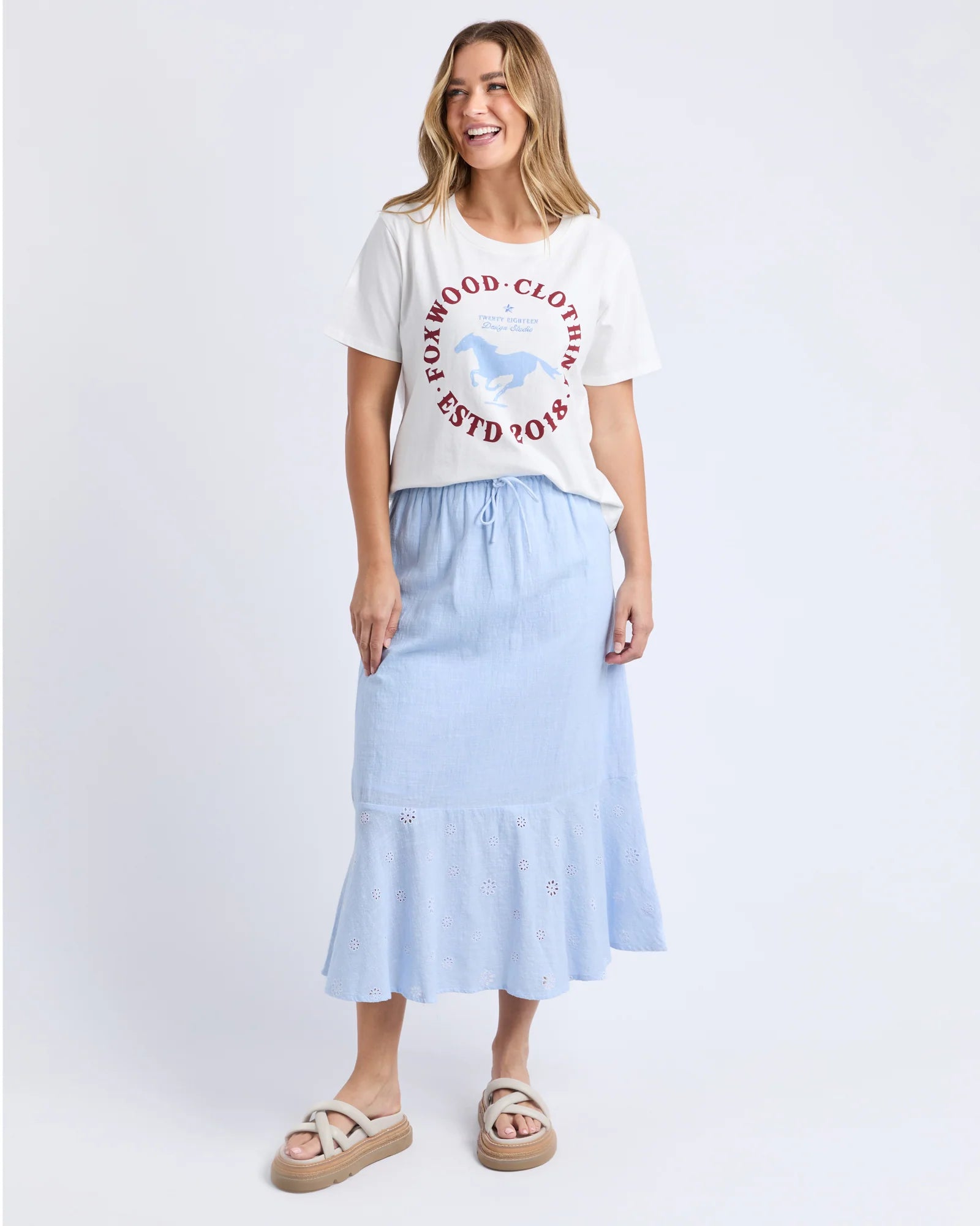 Image of A woman modeling a light blue midi skirt with a drawstring waist and ruffled hem, paired with a Foxwood white graphic t-shirt and beige slide sandals, against a light gray studio background.