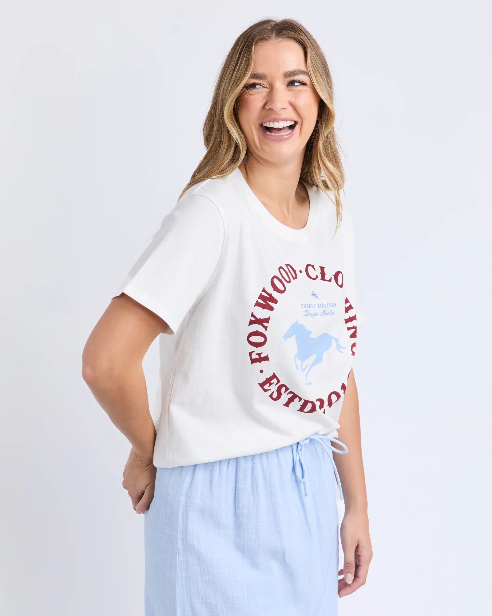 Image of A woman modeling a Foxwood white short-sleeve t-shirt with a red and blue circular graphic print that reads 'FOXWOOD CLOTHING ESTD 2015' and features a horse silhouette. She is wearing light blue drawstring pants against a clean white studio background.