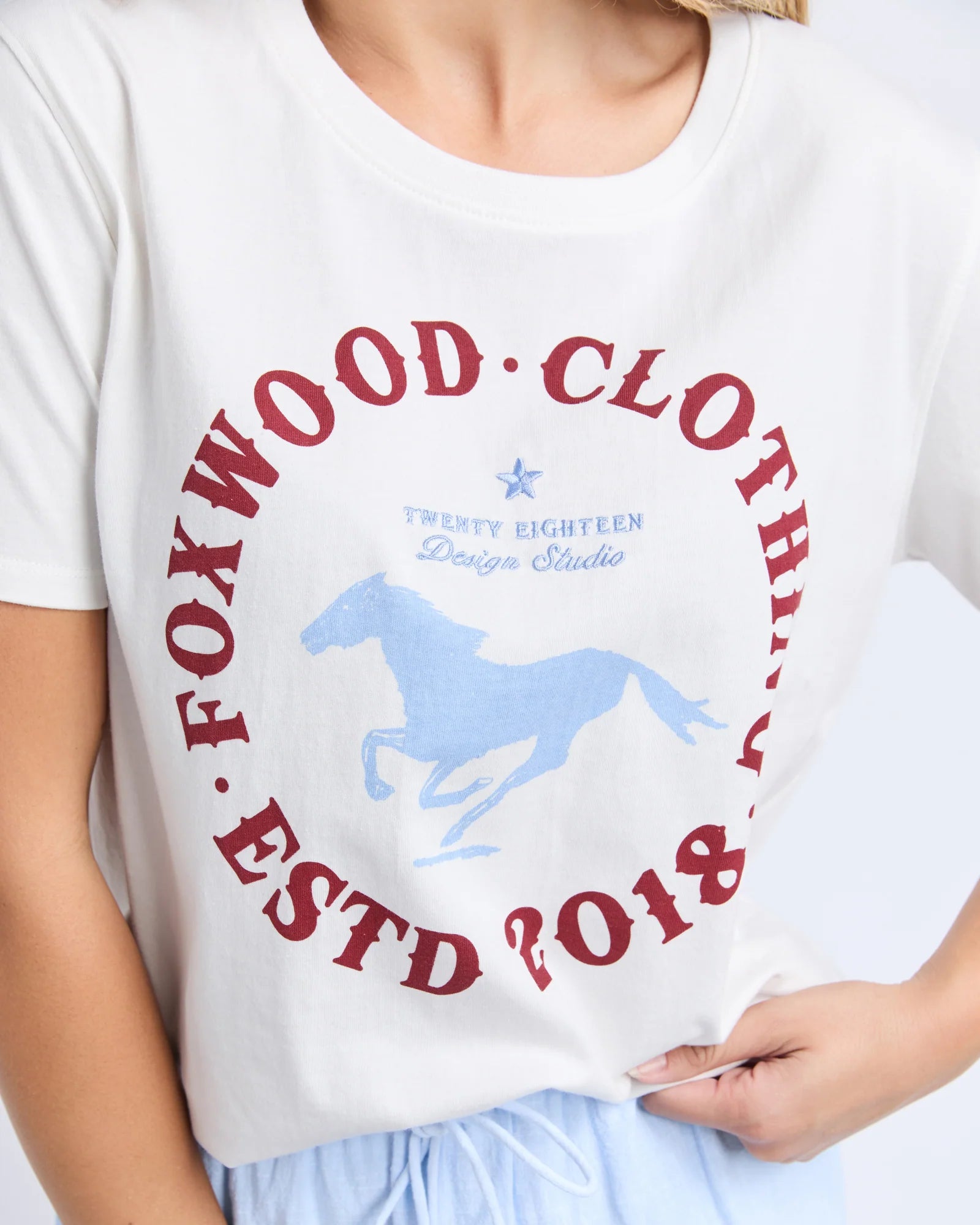 Image of A close-up of a woman wearing a Foxwood white short-sleeve graphic T-shirt. The shirt features a large circular red text print reading 'FOXWOOD CLOTHING ESTD 2018' surrounding a light blue horse silhouette and smaller blue text reading 'TWENTY EIGHTEEN Design Studio'.