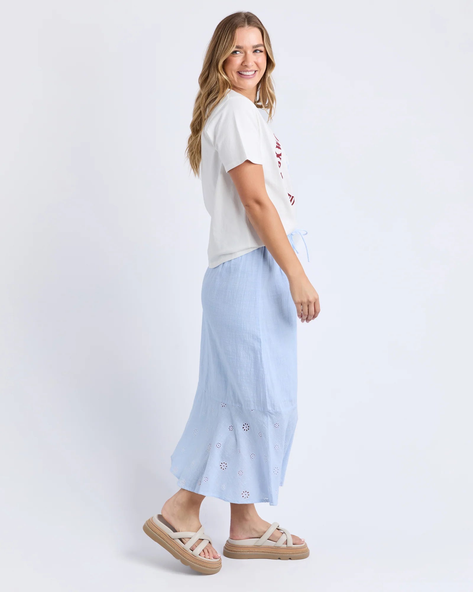 Image of A woman modeling a light blue midi skirt with eyelet embroidery, paired with a Foxwood white t-shirt and beige platform sandals, against a plain white studio background.