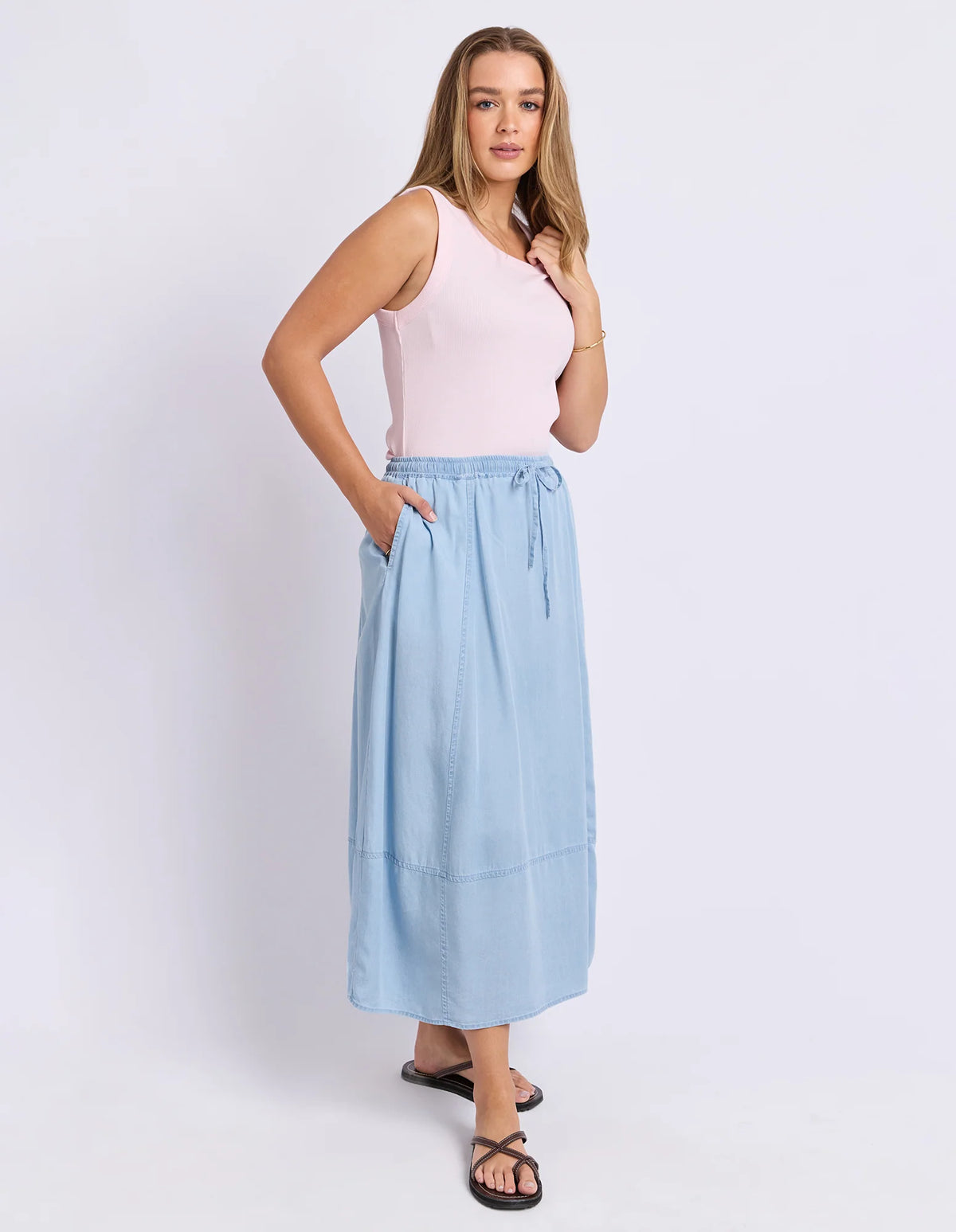 A woman modeling a Foxwood light blue midi skirt with a drawstring waist and side pockets, paired with a pale pink tank top and black sandals, against a plain white studio background.