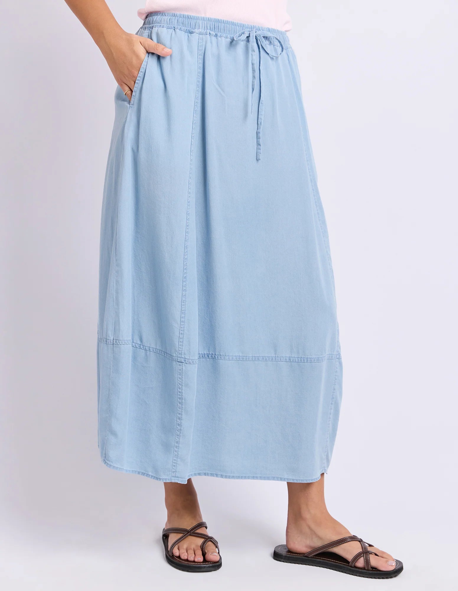 Image of A woman wearing a Foxwood light blue midi skirt with side pockets and a drawstring waist, paired with black strappy sandals against a plain light gray studio background.