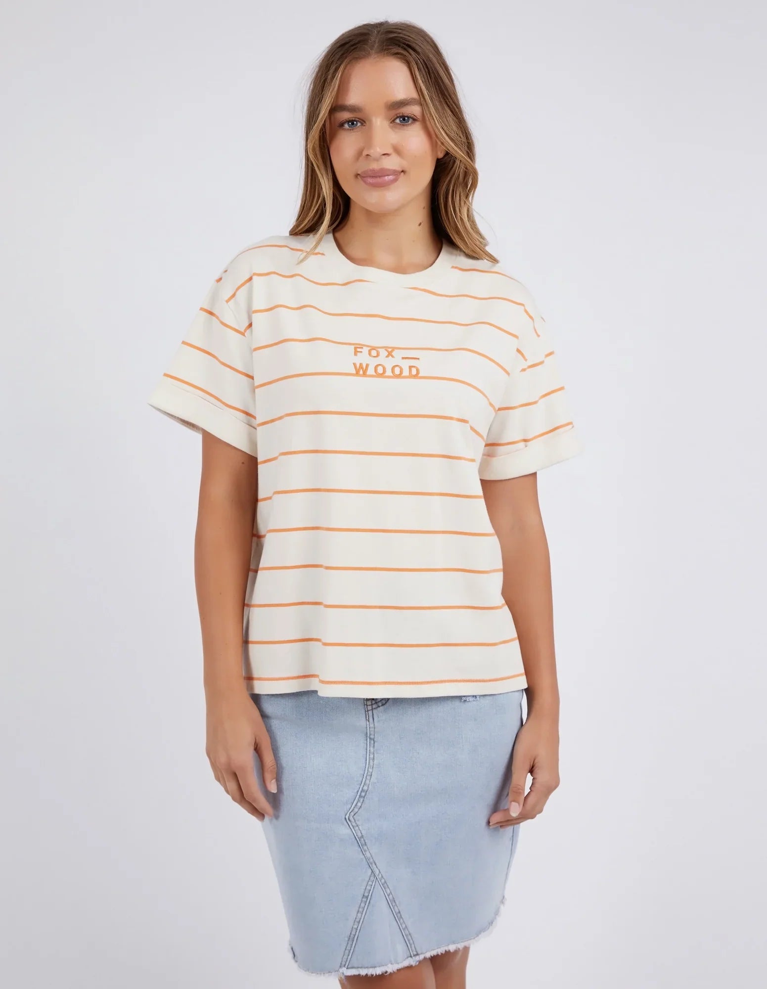 Image of A woman modeling a Foxwood short-sleeve crewneck t-shirt with thin orange horizontal stripes on a cream base, featuring the text 'fox wood' printed in orange on the chest. She is also wearing a light blue denim skirt, set against a plain light gray studio background.