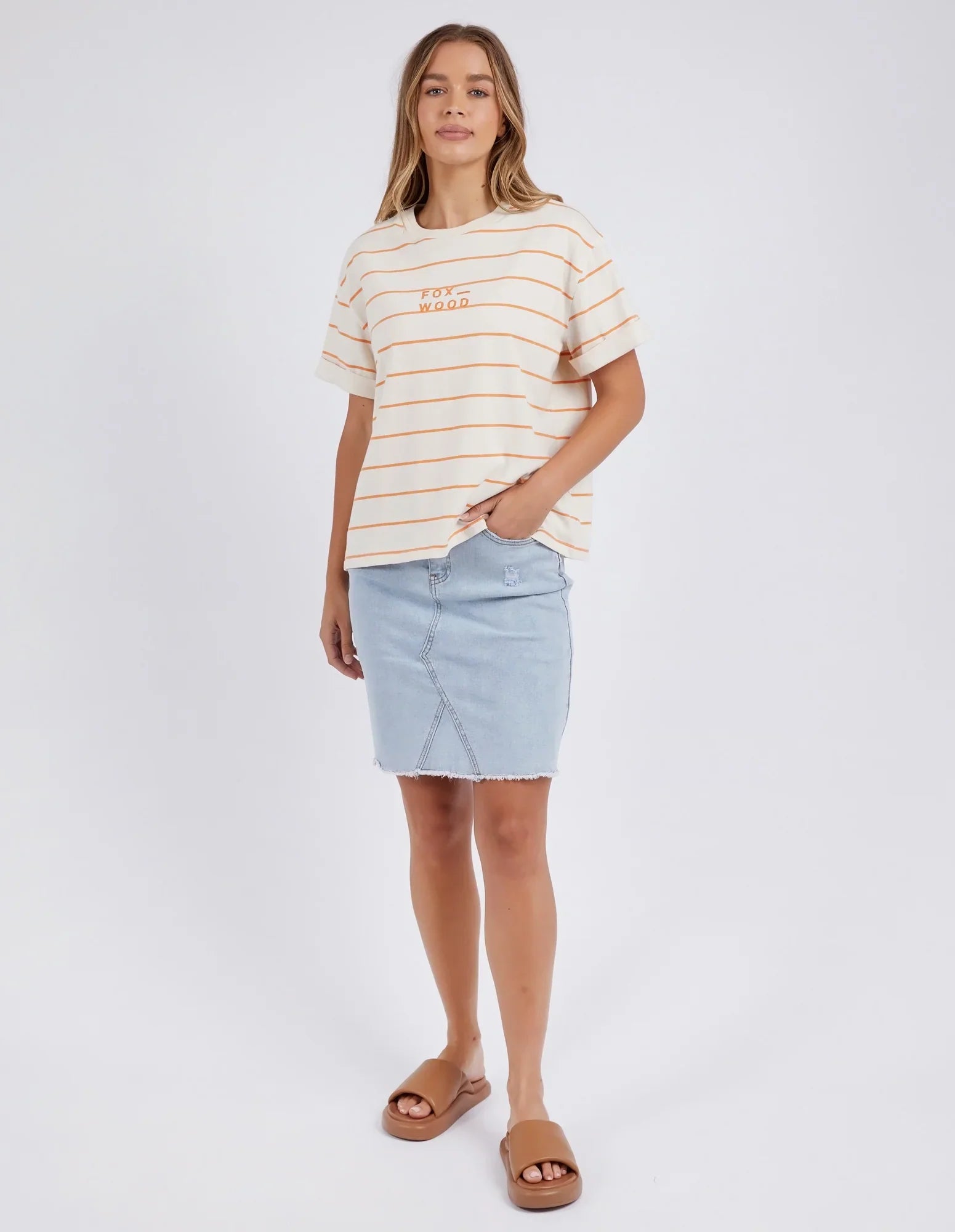 Image of A woman modeling a Foxwood short-sleeve crewneck t-shirt with thin orange stripes on an off-white base, featuring a small orange graphic on the left chest. She is wearing a light-wash denim mini skirt and brown slide sandals, standing against a plain light gray studio background.