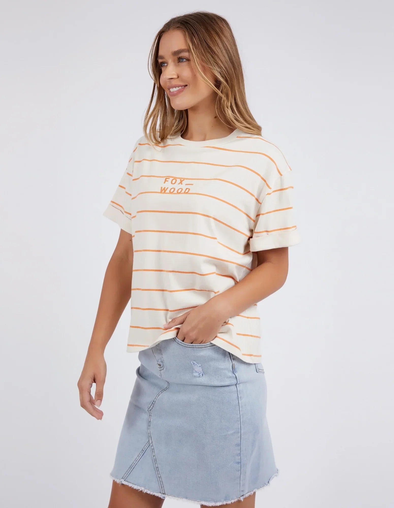 A woman modeling a Foxwood cream-colored short-sleeve t-shirt with thin orange horizontal stripes and 'FOX WOOD' embroidered on the chest, paired with a light blue denim mini skirt, against a plain light gray background.
