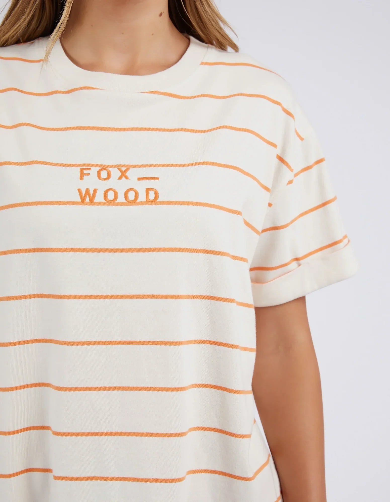 Image of A close-up of a woman wearing a Foxwood short-sleeve crew-neck T-shirt with thin orange horizontal stripes on an off-white base, featuring the text 'FOX WOOD' printed in orange on the chest.