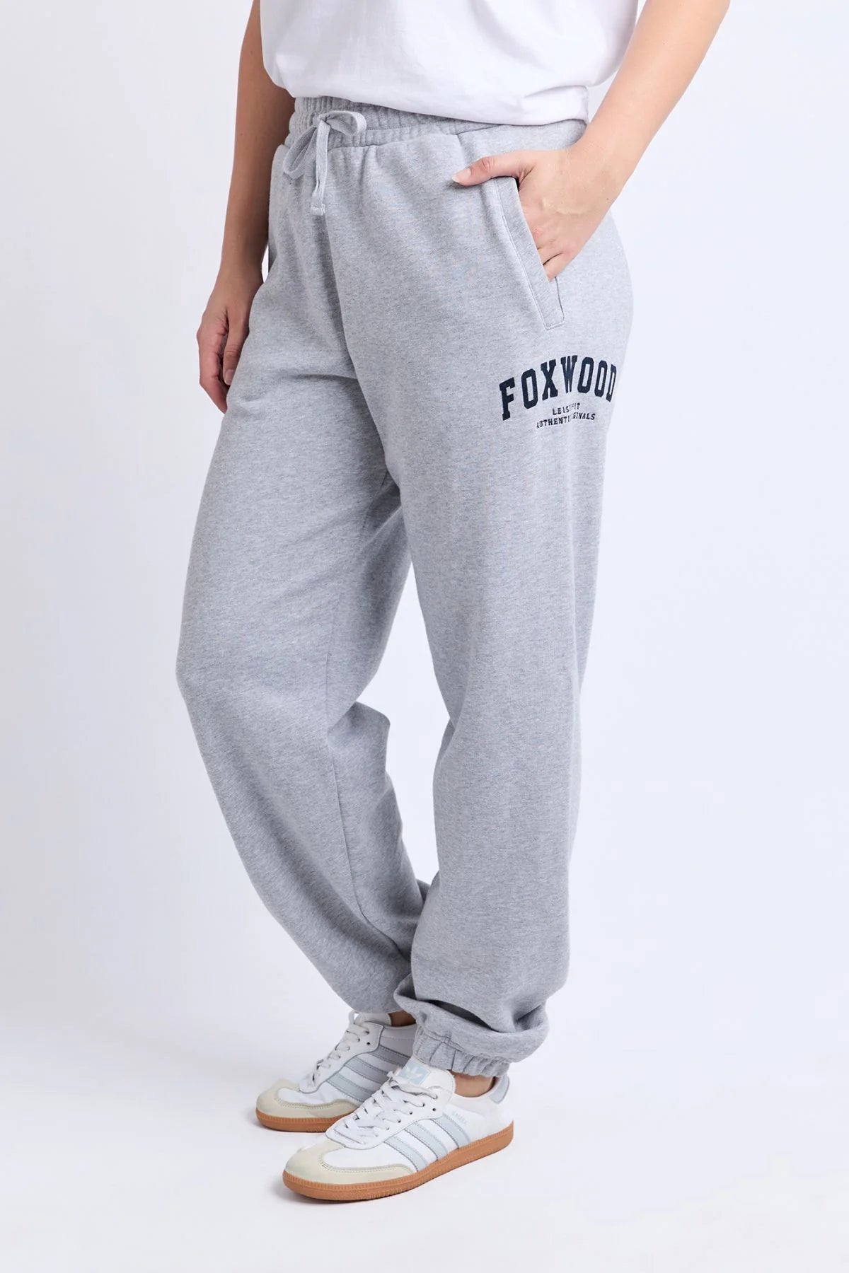 Image of Woman wearing grey Foxwood trackpants with a white top on a plain background 