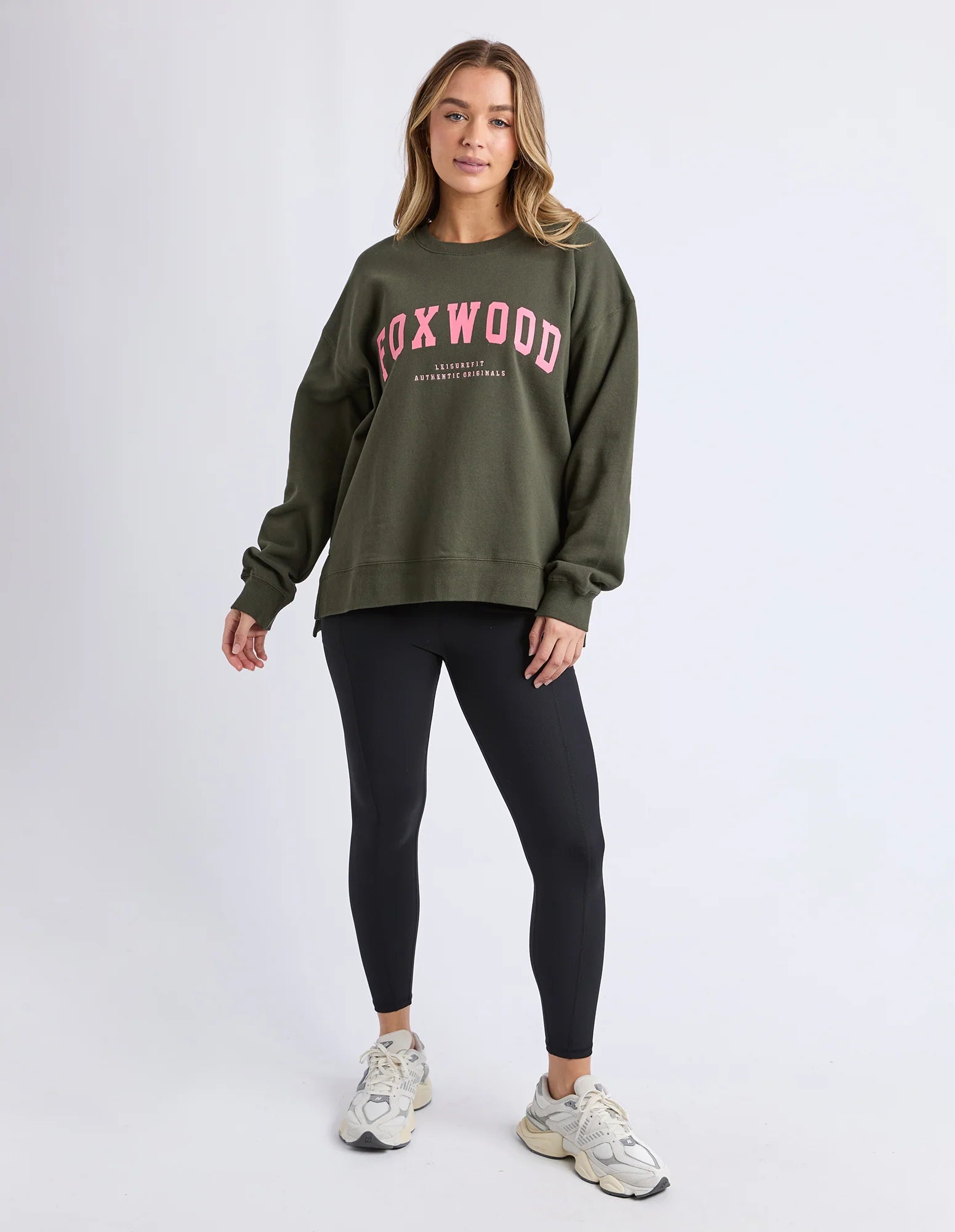 Woman wearing an olive green sweatshirt with 'FOXWOOD' printed on it, black leggings, and white sneakers against a white background.