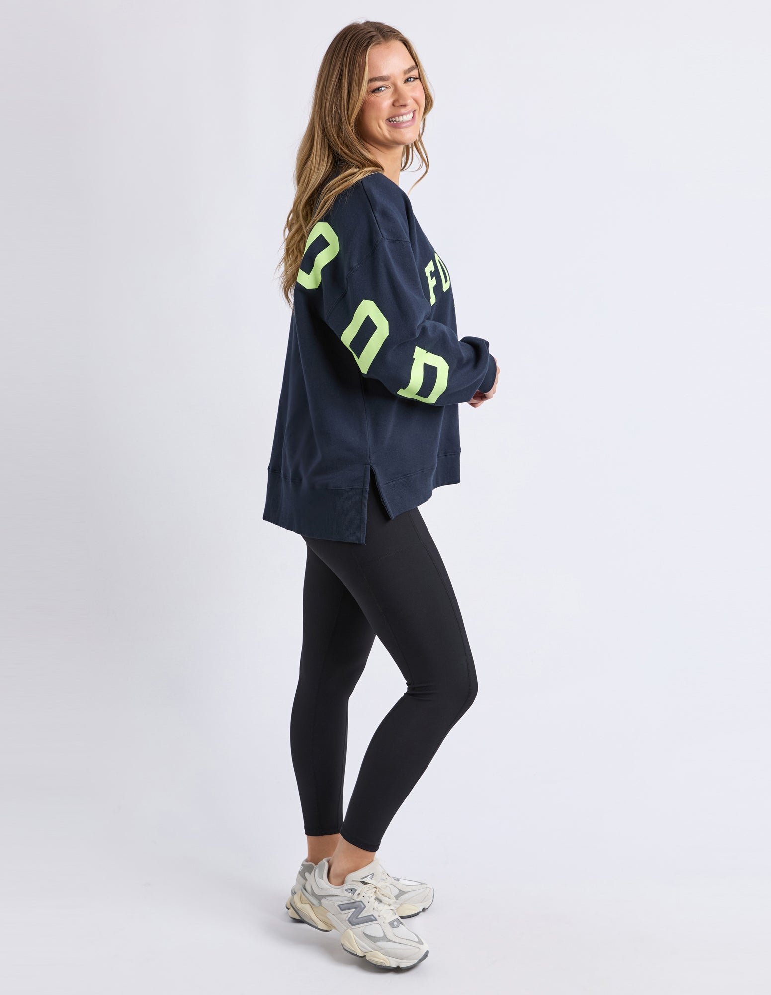 Image of Woman wearing a Foxwood navy sweater with bright green letters and black leggings on a white background
