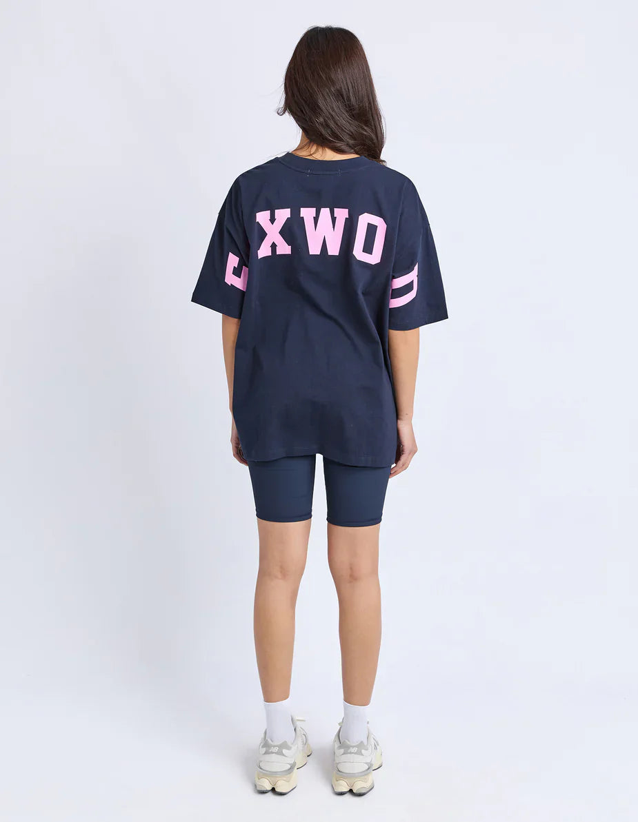 Image of Woman wearing a navy blue oversized t-shirt with 'FOXWOOD' printed in pink on the back, standing against a white background.
