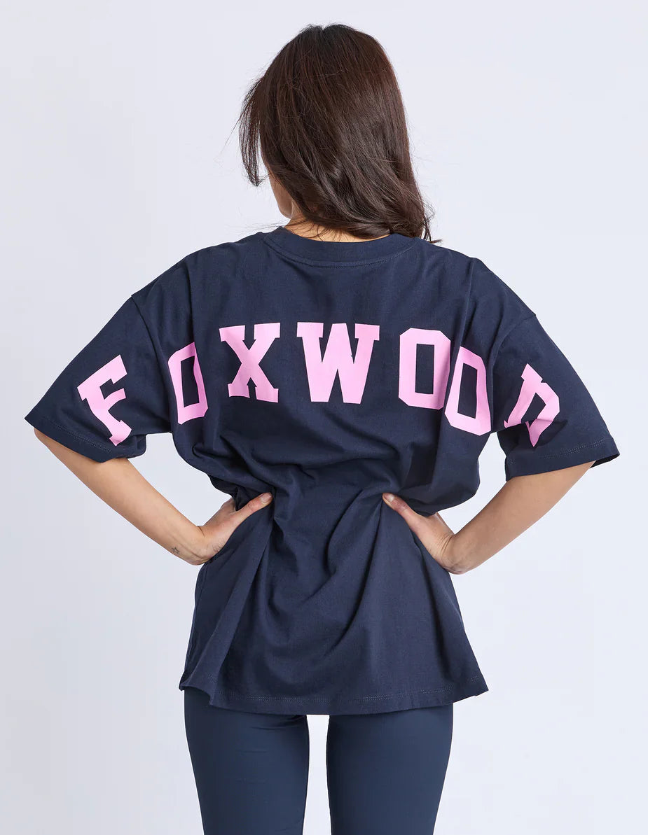 Woman wearing a Foxwood navy blue shirt with 'FOXWOOD' printed in pink on the back against a white background