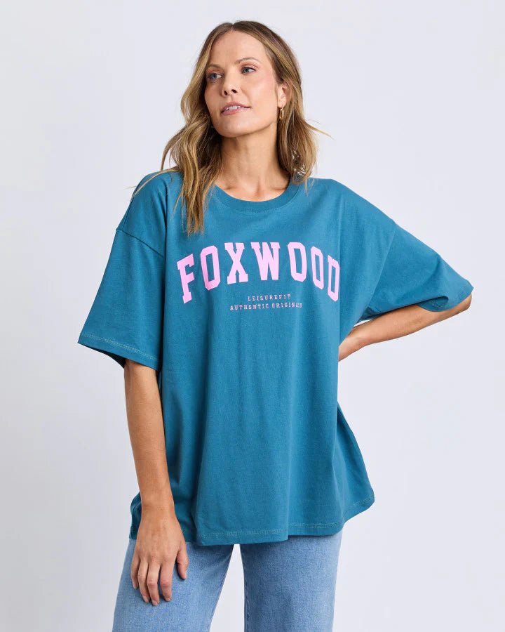 Image of Woman wearing a Foxwood teal t-shirt with 'FOXWOOD' printed in pink on a light gray background