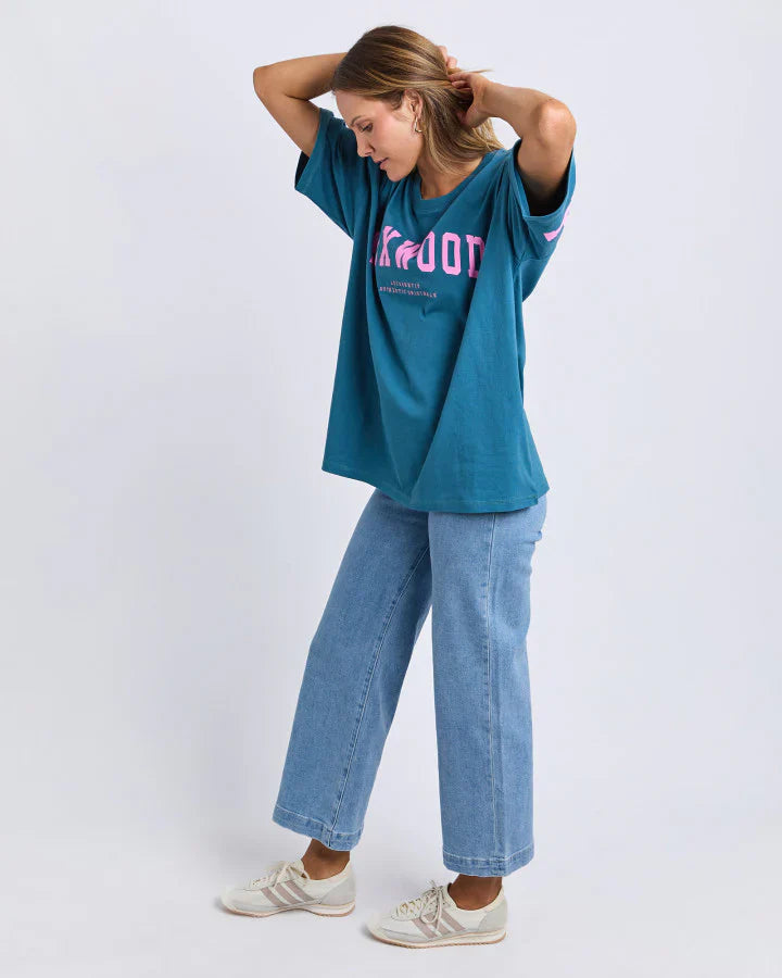 Woman wearing a Foxwood teal oversized t-shirt with pink text and light blue jeans on a white background