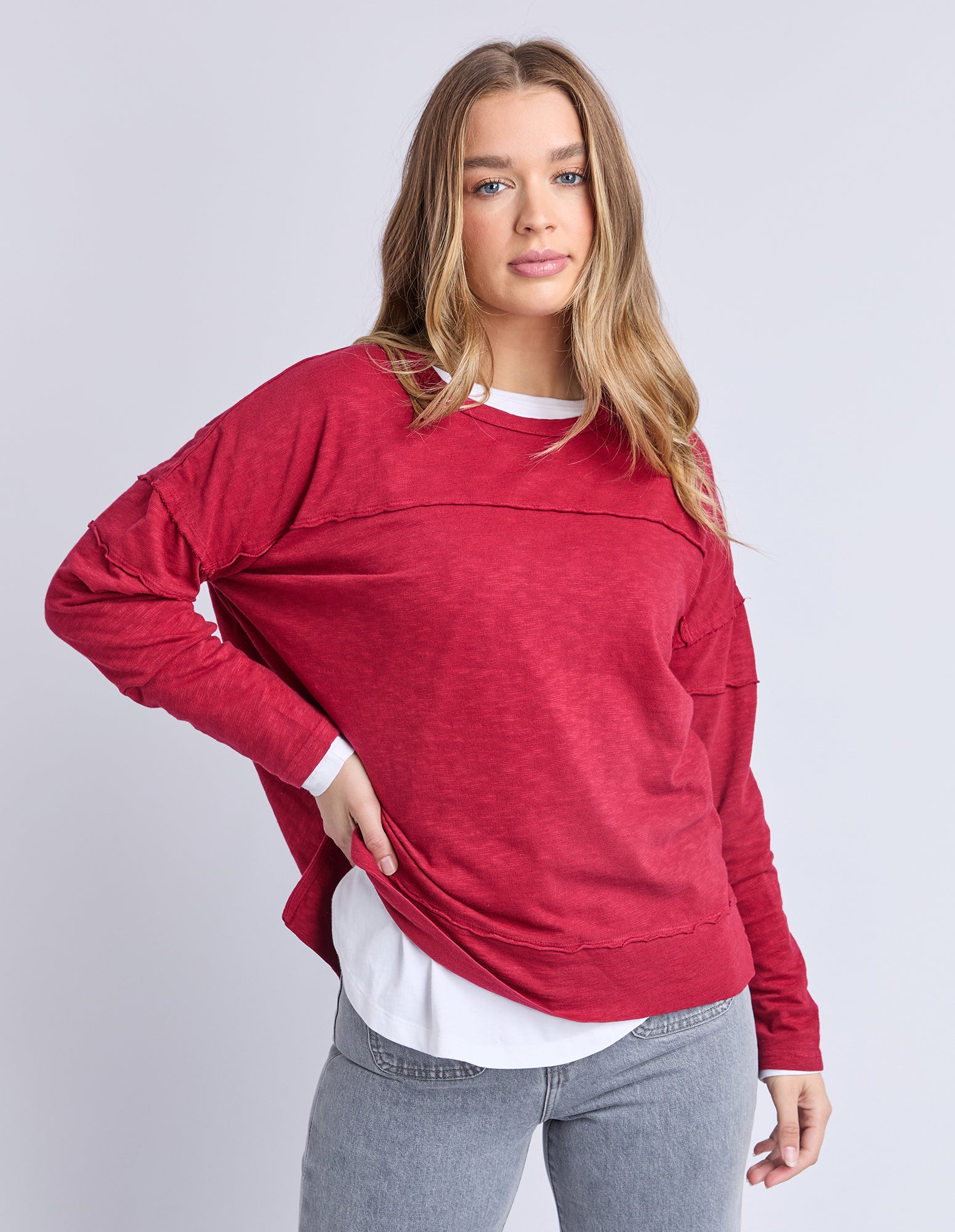 Woman wearing a red long sleeve tee over a white shirt with grey pants on a light grey background