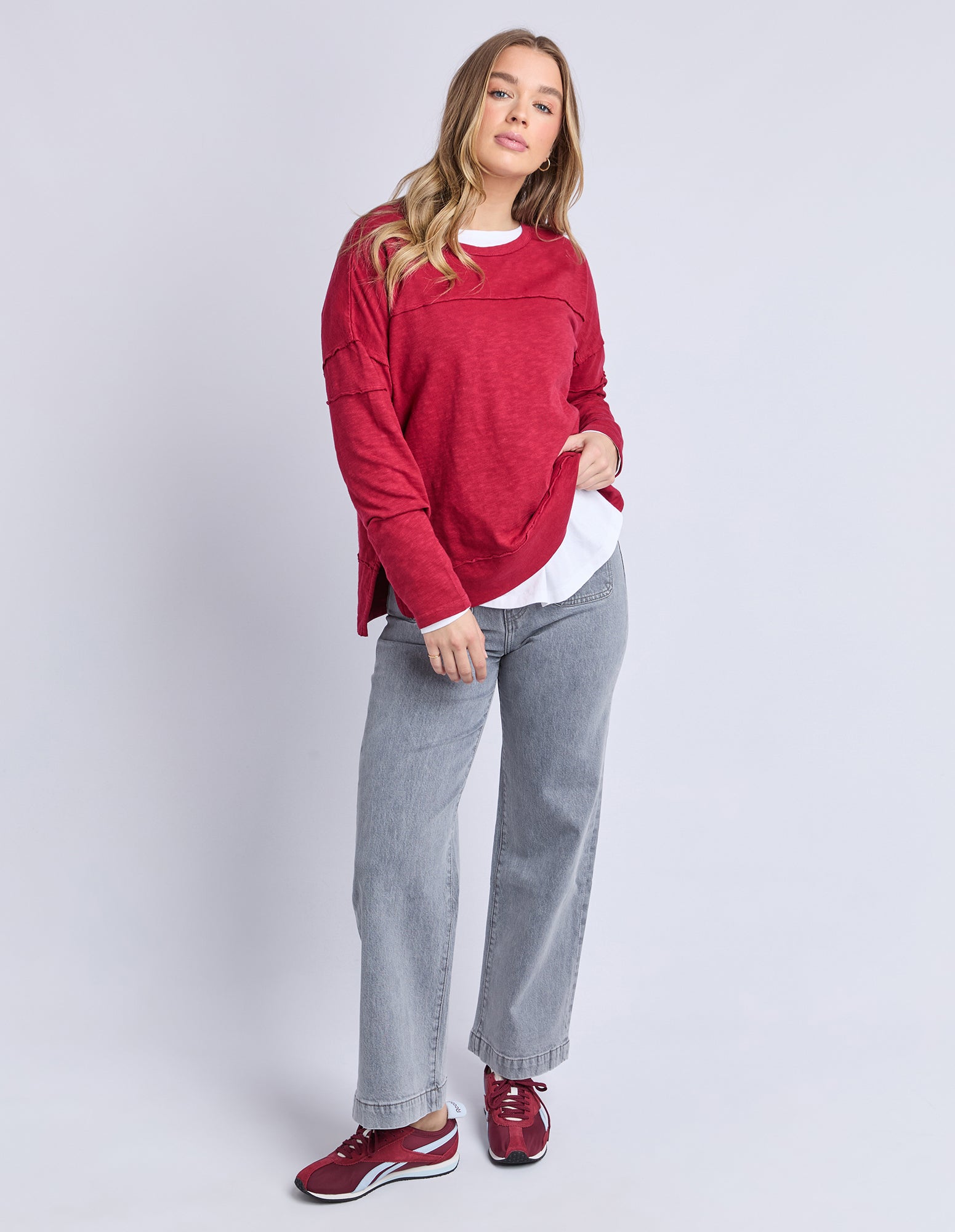 Image of Woman wearing a Foxwood red long sleeve top, white shirt, grey jeans, and red sneakers on a light grey background