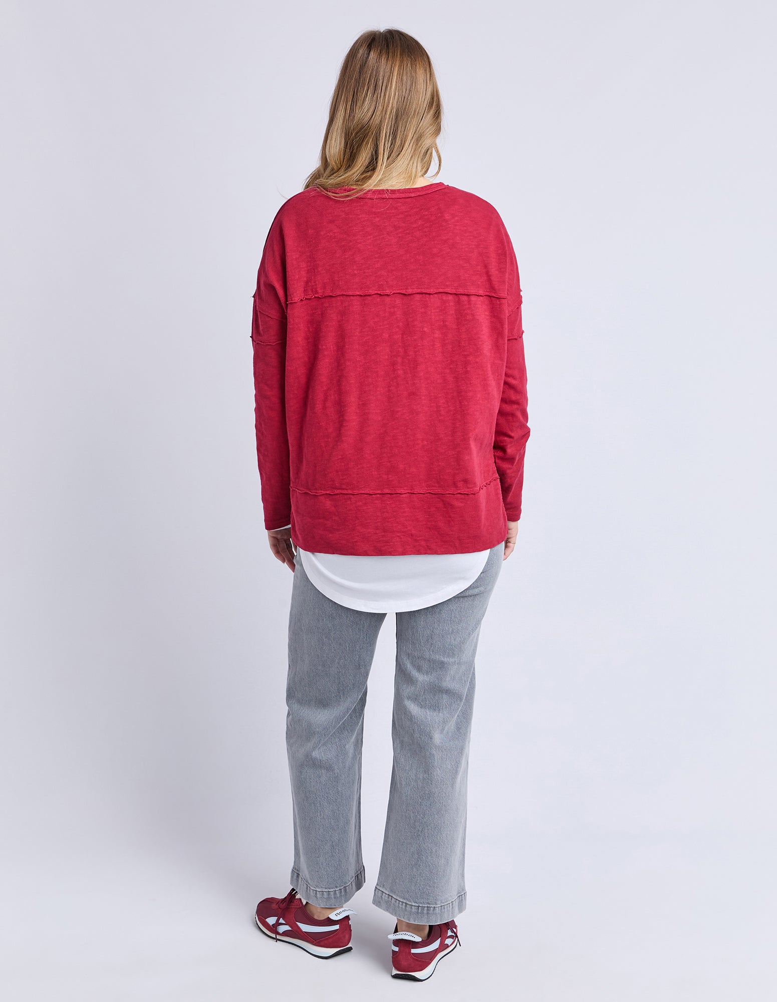 Back view of a woman wearing a Foxwood red long sleeve top, white shirt, grey pants, and red shoes on a light grey background