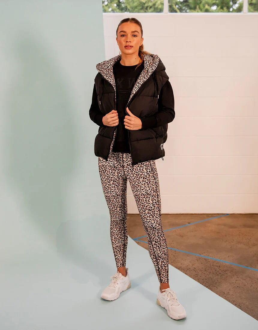 A woman modeling a black puffer vest with a leopard-print hood over a black top and matching Foxwood leopard-print leggings, paired with white sneakers, in an indoor studio setting.