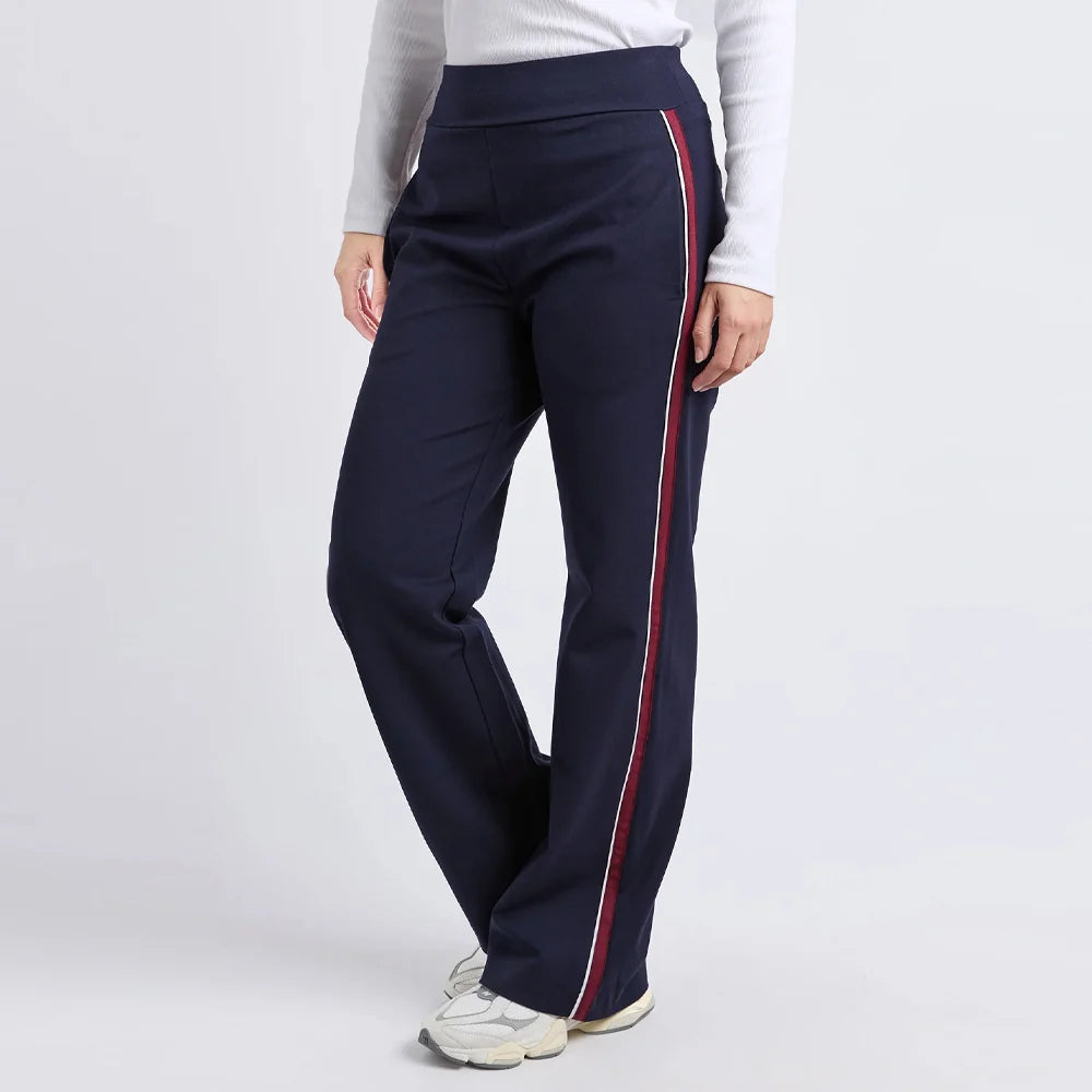 A woman wearing Foxwood navy wide-leg track pants with a red and white side stripe, paired with a white long-sleeve top and white sneakers, shown against a light gray studio background.