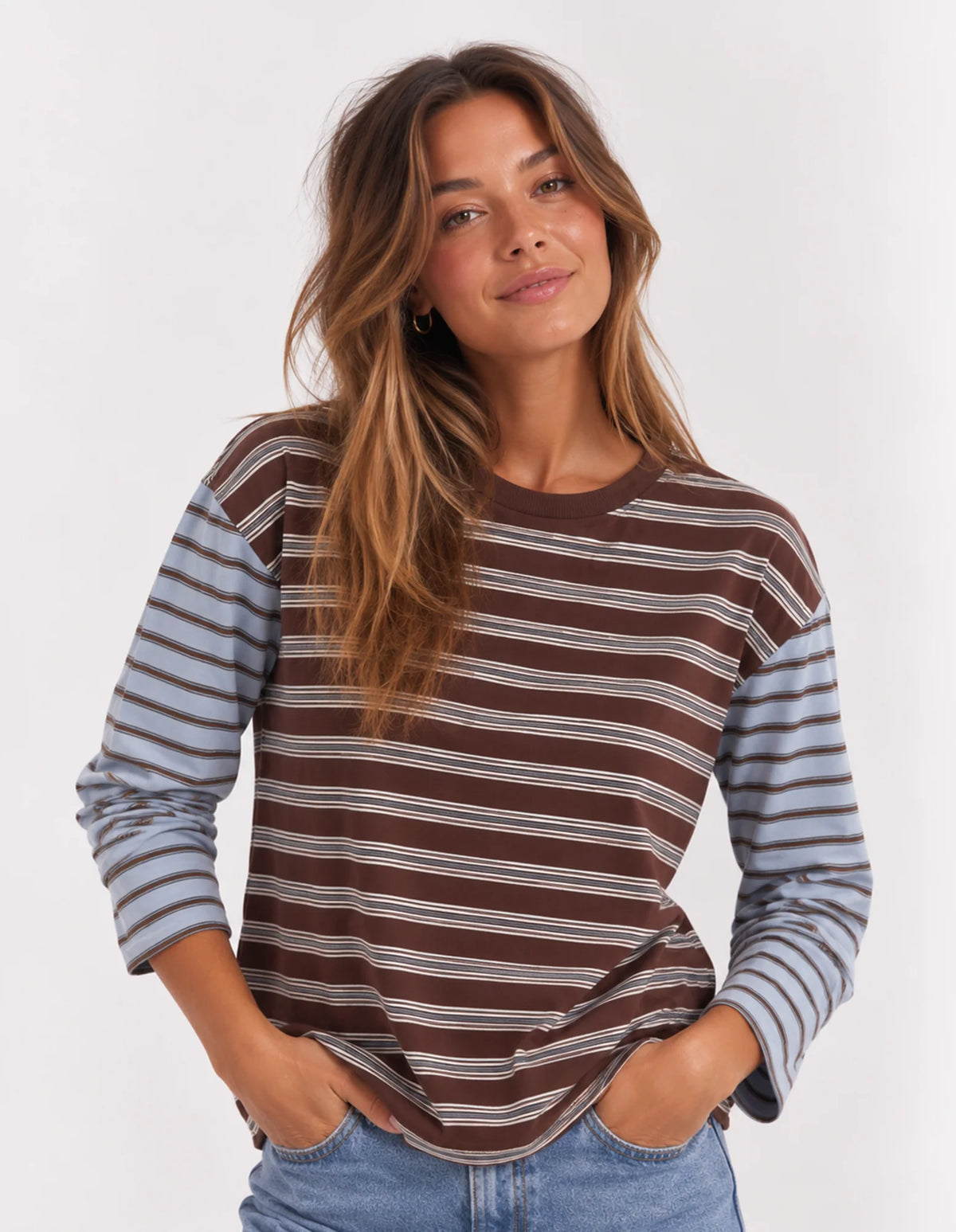 A woman wearing a Foxwood long-sleeve striped shirt with a brown body and muted blue-gray sleeves, paired with blue jeans, against a plain light background.