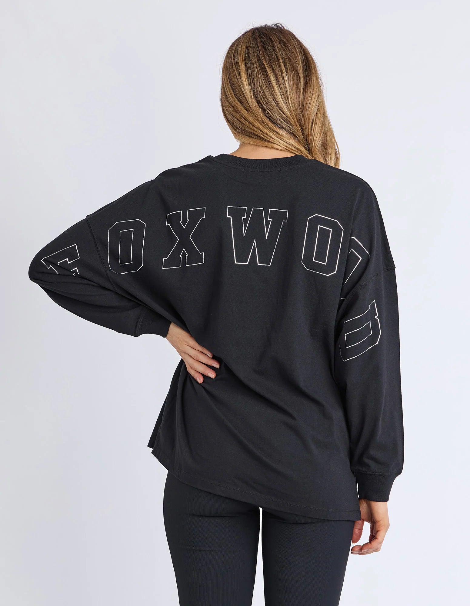 Image of Woman wearing Foxwood long sleeve top with Foxwood branding pictured on the front and back of top. Top paired with black leggings on a plain background