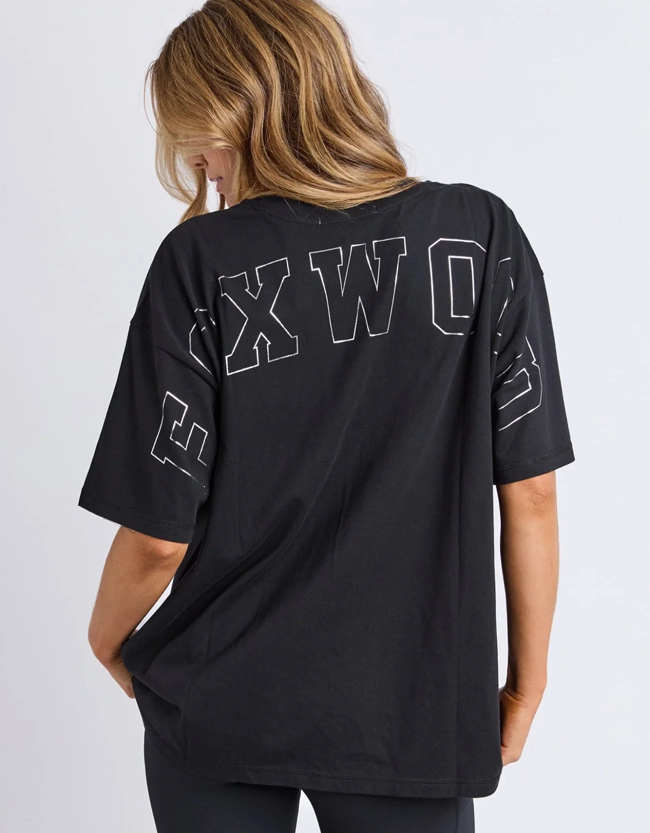 Woman wearing a Foxwood black oversized t-shirt with white text on the back against a plain background
