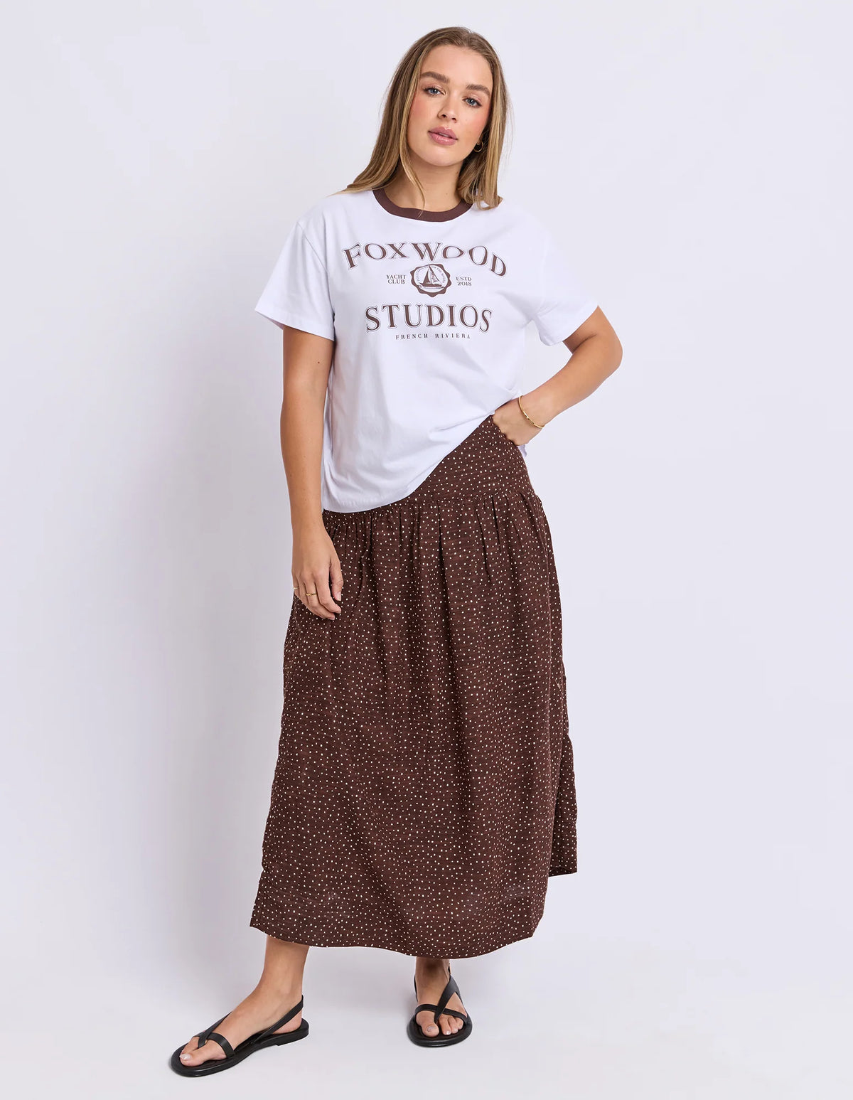 A woman modeling a white Foxwood Studios graphic t-shirt and a brown patterned midi skirt, paired with black flat sandals, against a light gray studio background.