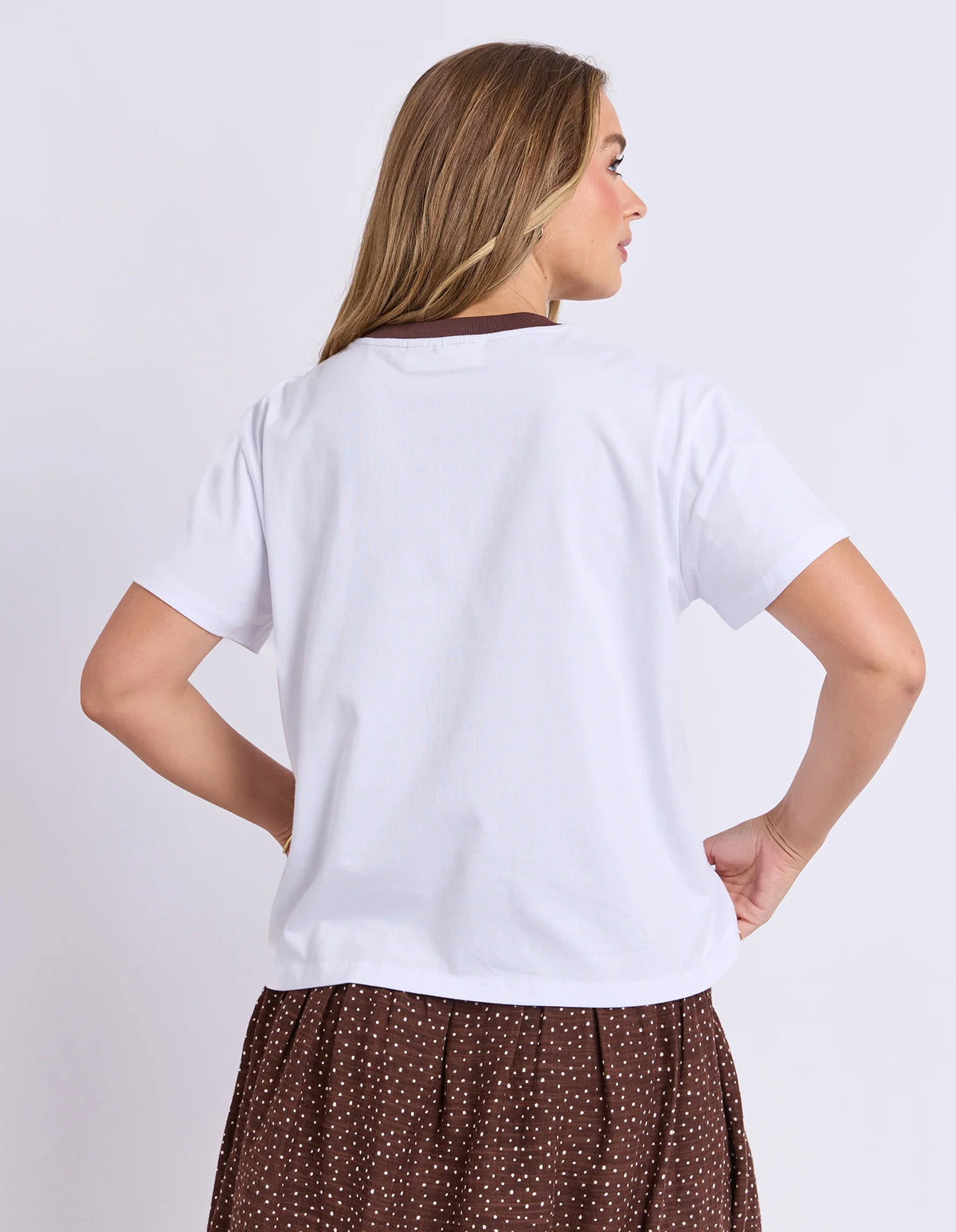 A woman shown from the back wearing a Foxwood plain white short-sleeve t-shirt with a dark brown collar trim and a brown polka dot skirt, against a light gray studio background.