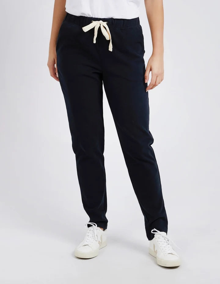 A woman standing in a studio setting, wearing Foxwood black cotton pants with an elastic waist and contrast drawcord, paired with a white top and white sneakers.