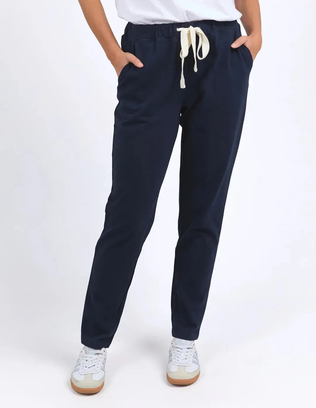 Image of Woman wearing navy trackpants paired with a white top on a plain background