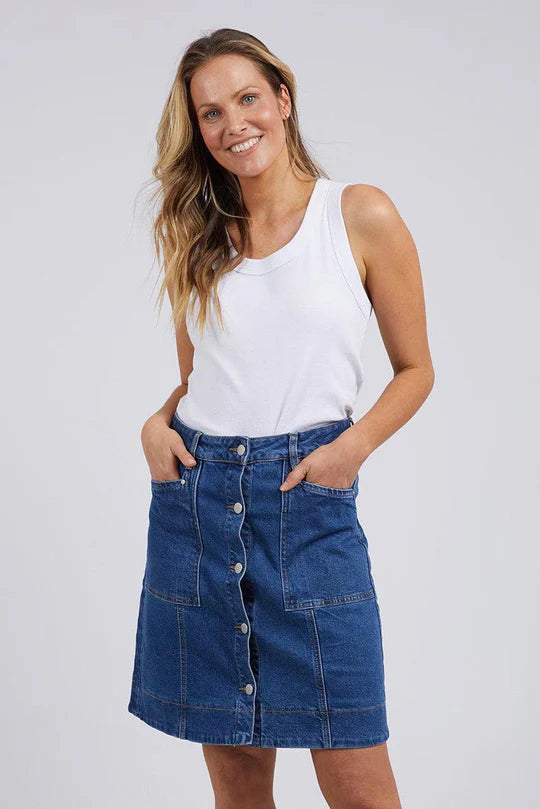 Image of A woman modeling a Foxwood blue denim button-front mini skirt with front pockets, paired with a white tank top, against a light gray studio background.