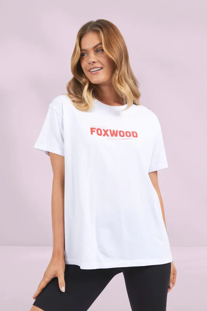 Image of A woman modeling an oversized Foxwood white t-shirt with 'FOXWOOD' printed in red across the chest, paired with black leggings, against a solid light pink studio background.