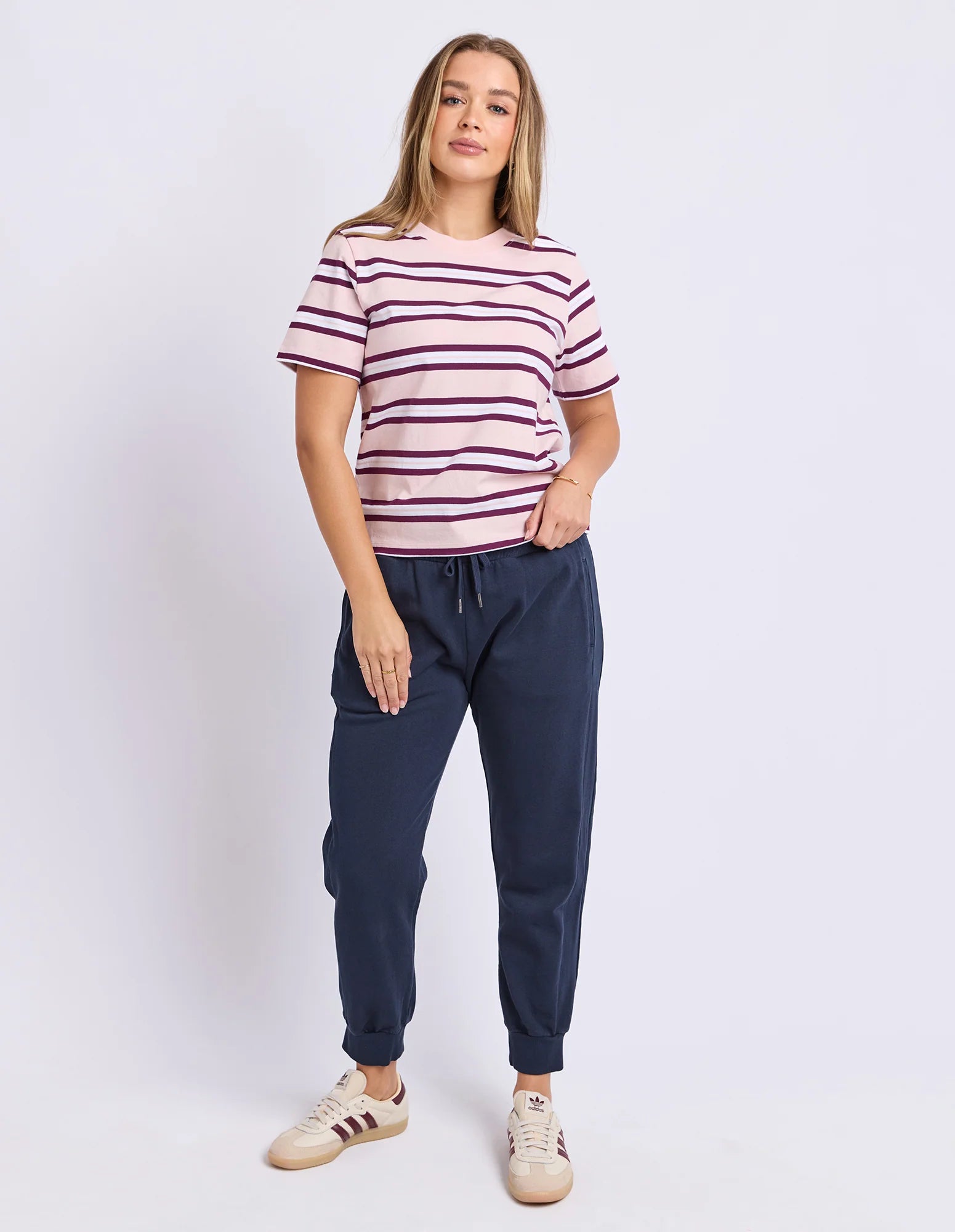 Image of A woman modeling a Foxwood short-sleeve crewneck t-shirt with horizontal pink and burgundy stripes, paired with navy blue jogger pants and beige sneakers, against a plain light gray studio background.