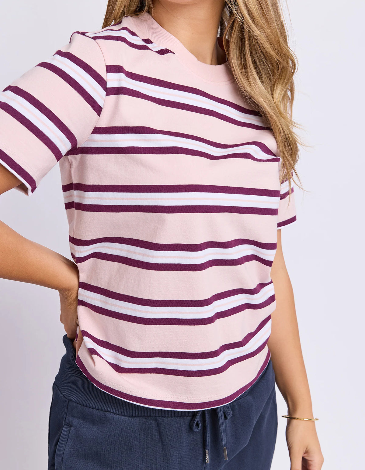 A woman wearing a Foxwood short-sleeve V-neck top with horizontal stripes in dusty pink, dark burgundy, and white, paired with navy drawstring pants against a light gray studio background.